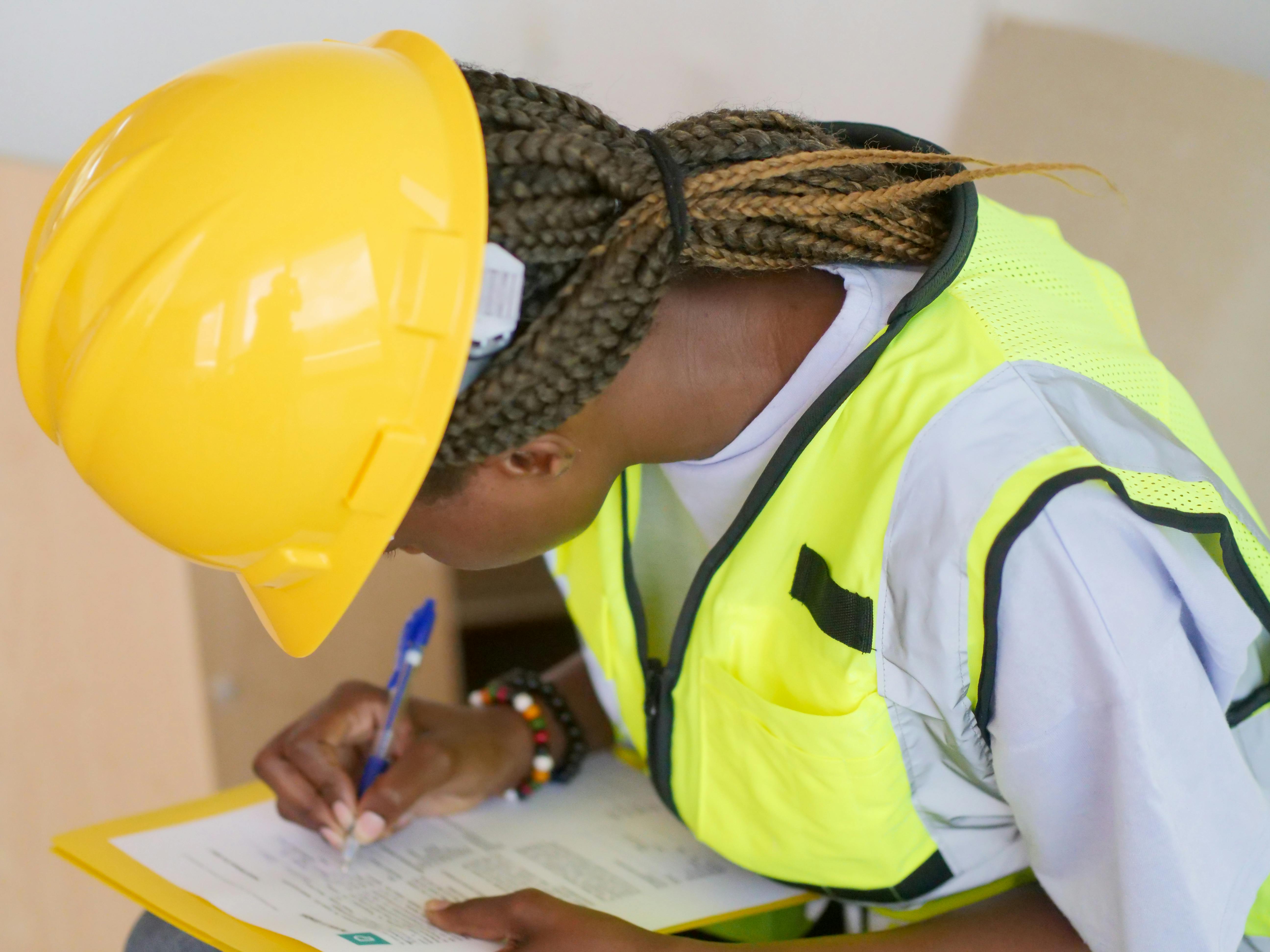 A dark-skinned woman with a braided ponytail wears a yellow hard hat, a yellow vest, and a gray tunic. She has a pen in her right hand and is bending over a form, completing it with the pen.