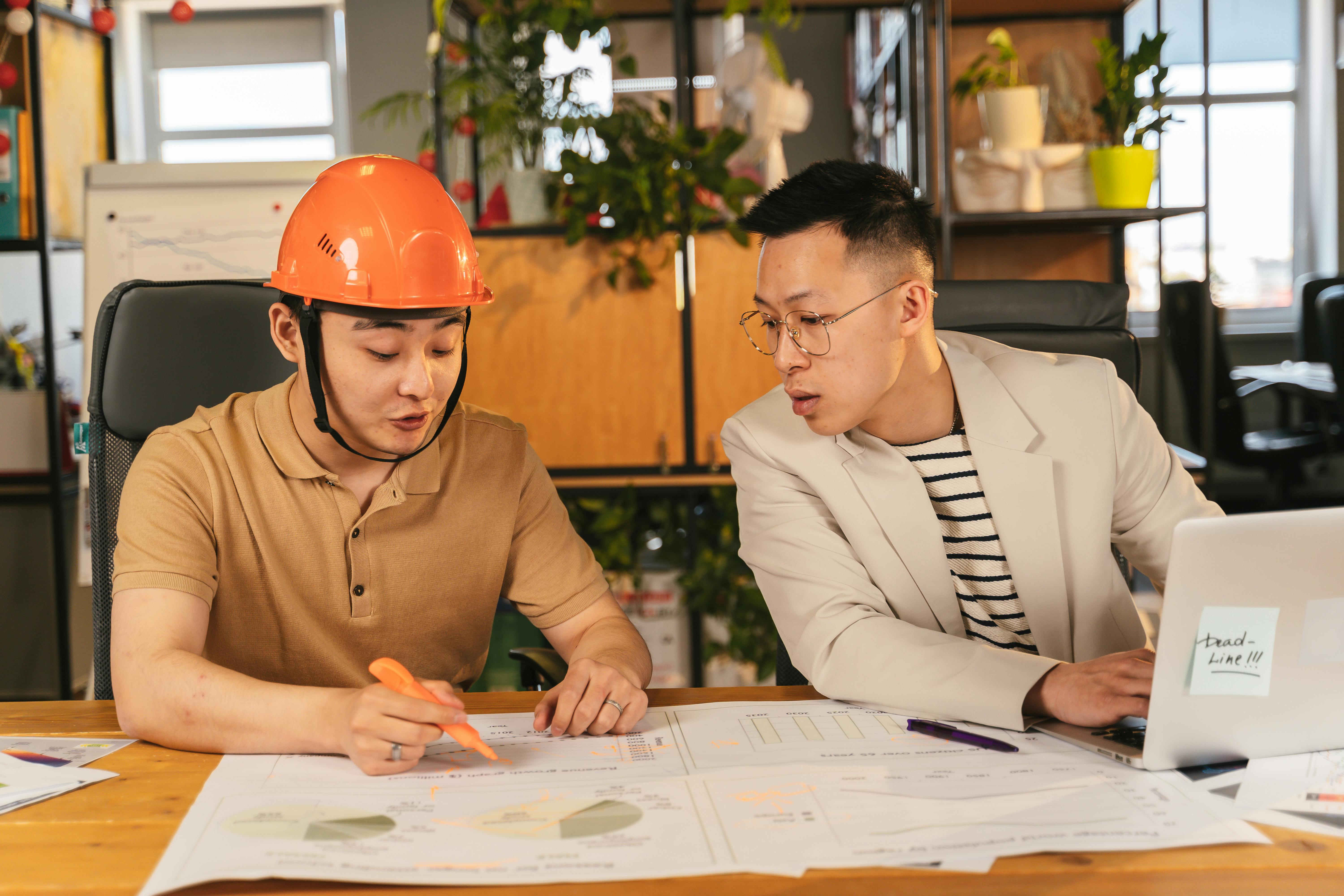 Two Asian men are sitting at a desk:
- One is wearing a t-shirt, holding an orange pen in his hand, and wearing an orange hard hat
- The gentleman next to him is wearing a suit jacket, glasses, and he's typing into a keyboard
- The gentleman on his right is pointing out things with an orange marker on what looks like a blueprint or a map