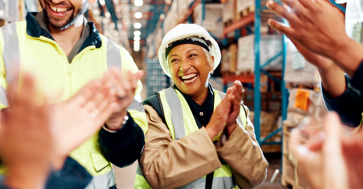 A photo of a group of workers in a warehouse all clapping and smiling, all wearing yellow vests. The woman in the center of the photograph, the most prominent, is absolutely beaming with glee.