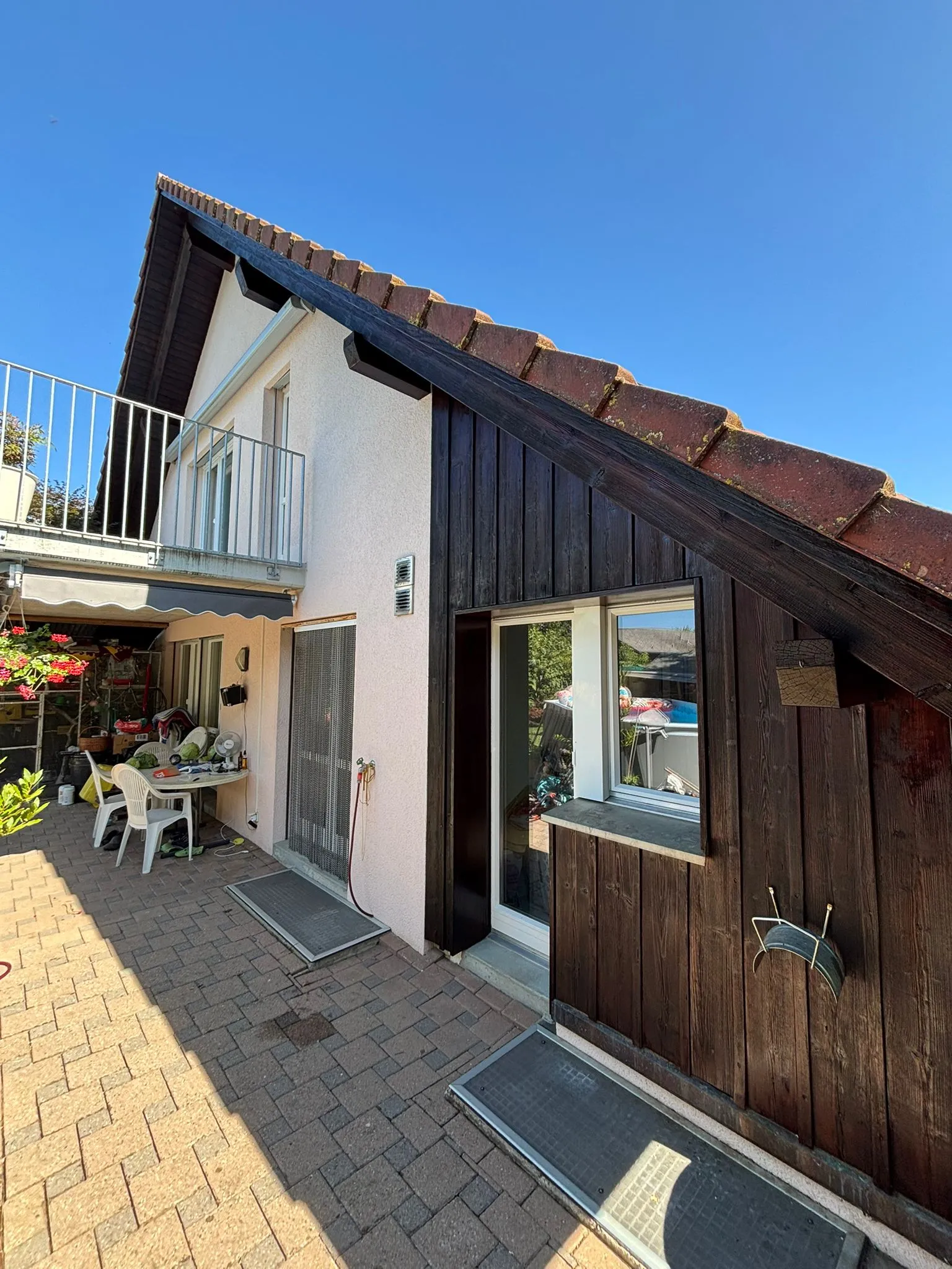 Haus mit einer Kombination aus weisser und dunkler Holzverkleidung, einem Balkon mit Geländer und einer gepflasterten Terrasse mit Stühlen und Pflanzen bei sonnigem Himmel.