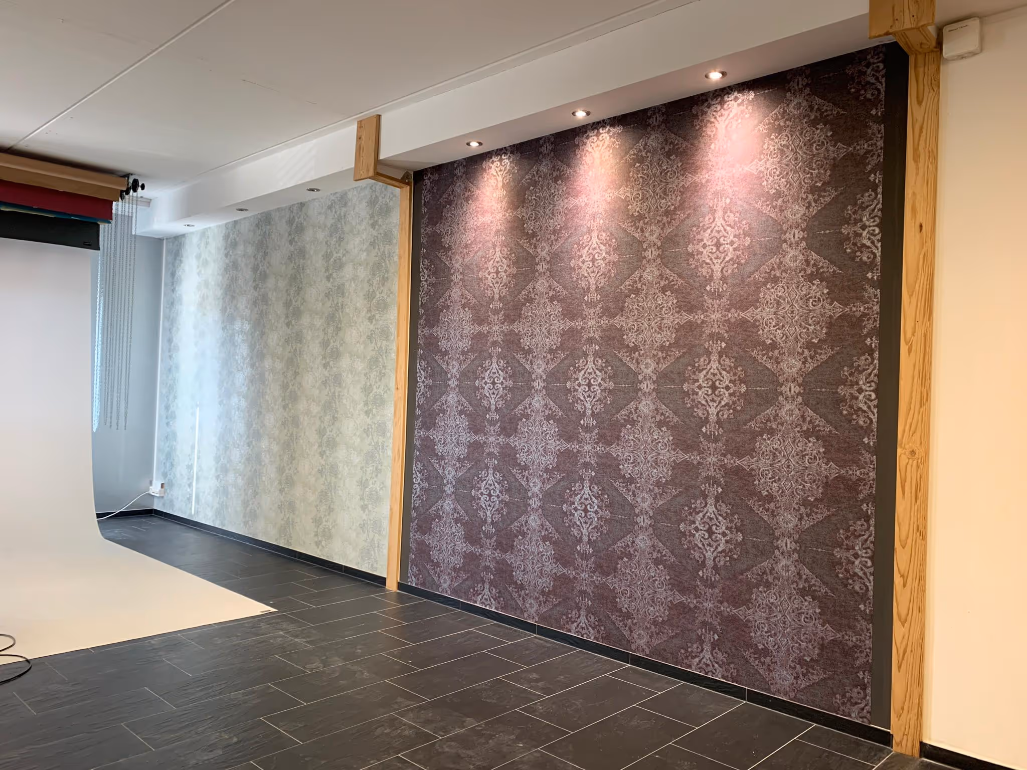 Innenraum mit zwei Wänden, eine mit hellem und eine mit dunklem gemustertem Tapetendesign, beleuchtet von Deckenstrahlern, und schwarzem gefliestem Boden.