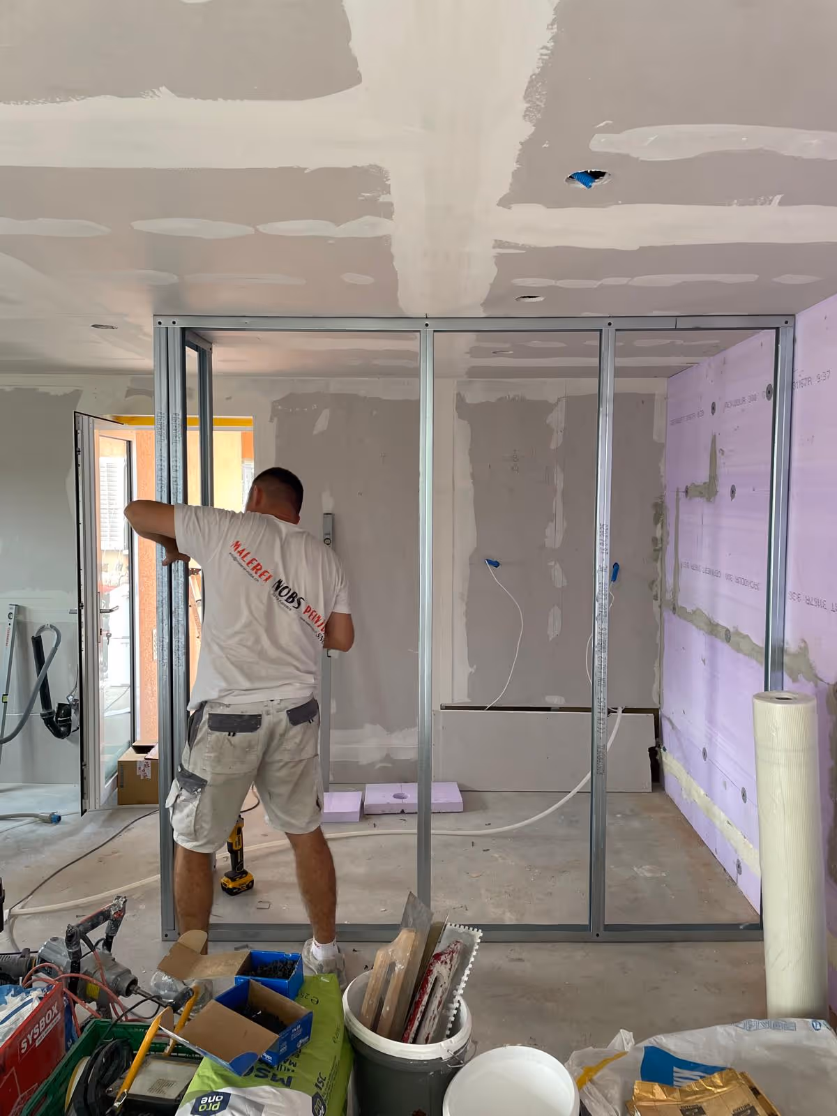 Handwerker montiert Metallrahmen für eine Wand in einem Rohbauzimmer mit Werkzeugen und Materialien auf dem Boden.