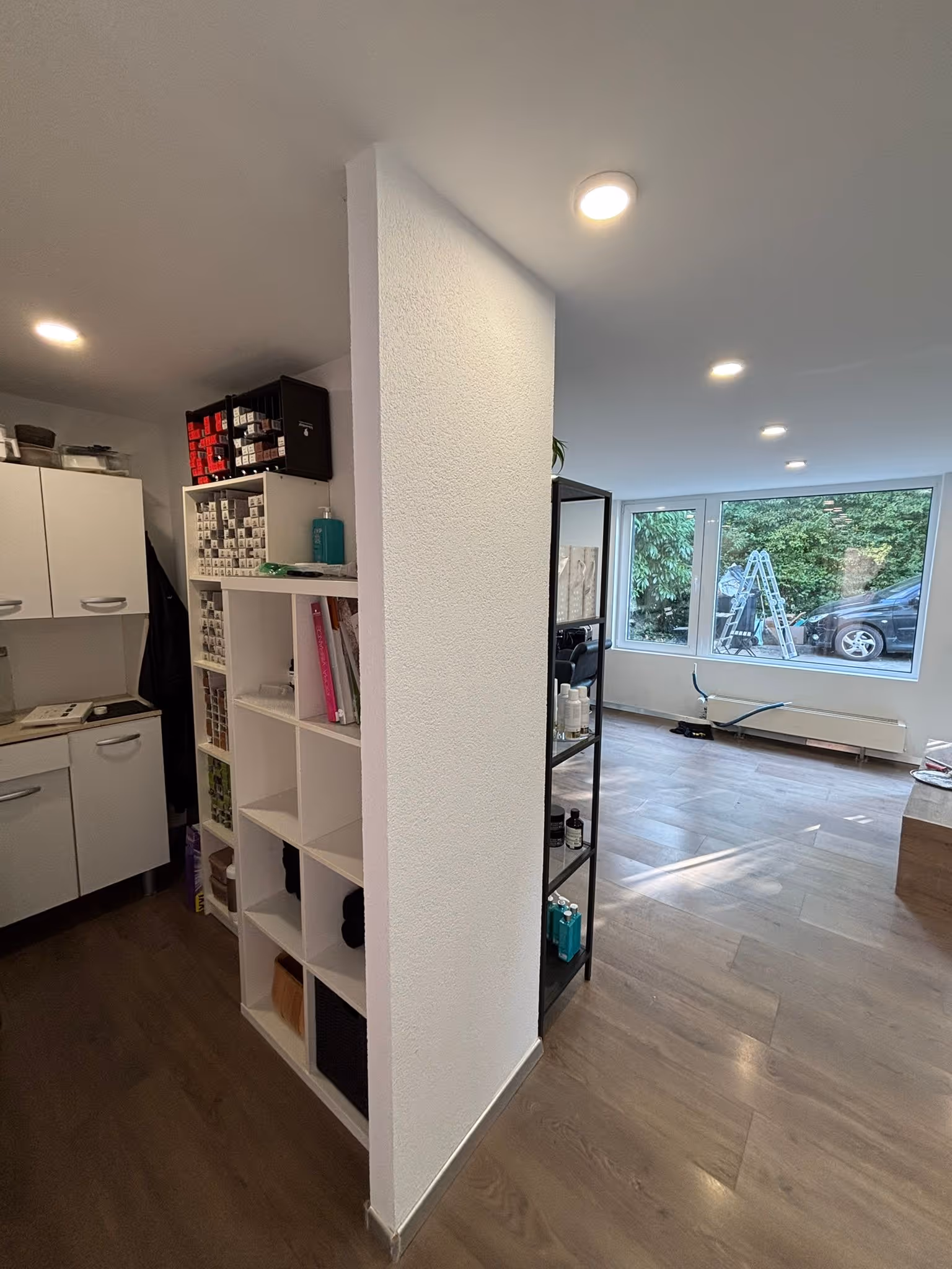 Innenraum mit weißer Wand in der Mitte, Regalen mit Produkten links und rechts, großen Fenstern mit Blick auf eine Autoleiter und grünes Laub draußen.