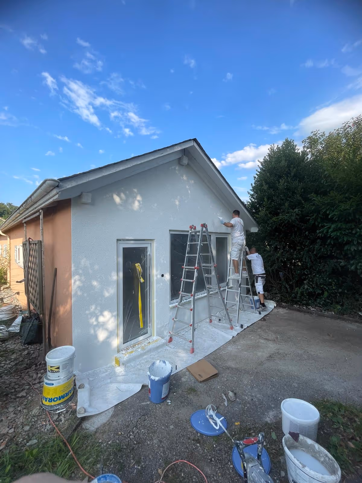 Zwei Männer streichen eine Hausfassade weiss, einer steht auf einer Leiter, die andere auf einer Trittleiter, davor Farbeimer und Malerwerkzeuge.