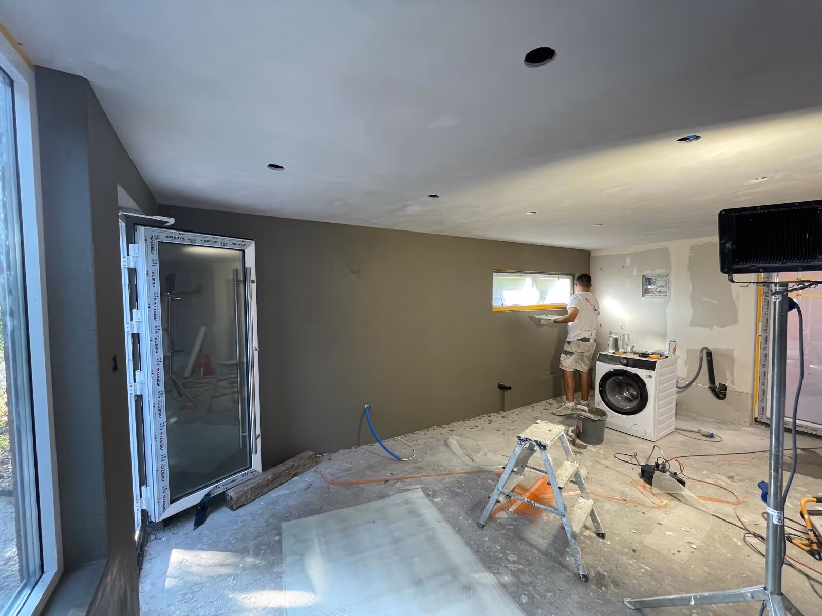 Zimmer in Renovierung mit offenem Fenster, einem Mann der eine Wand verputzt, einer Waschmaschine und Baustellenausrüstung.