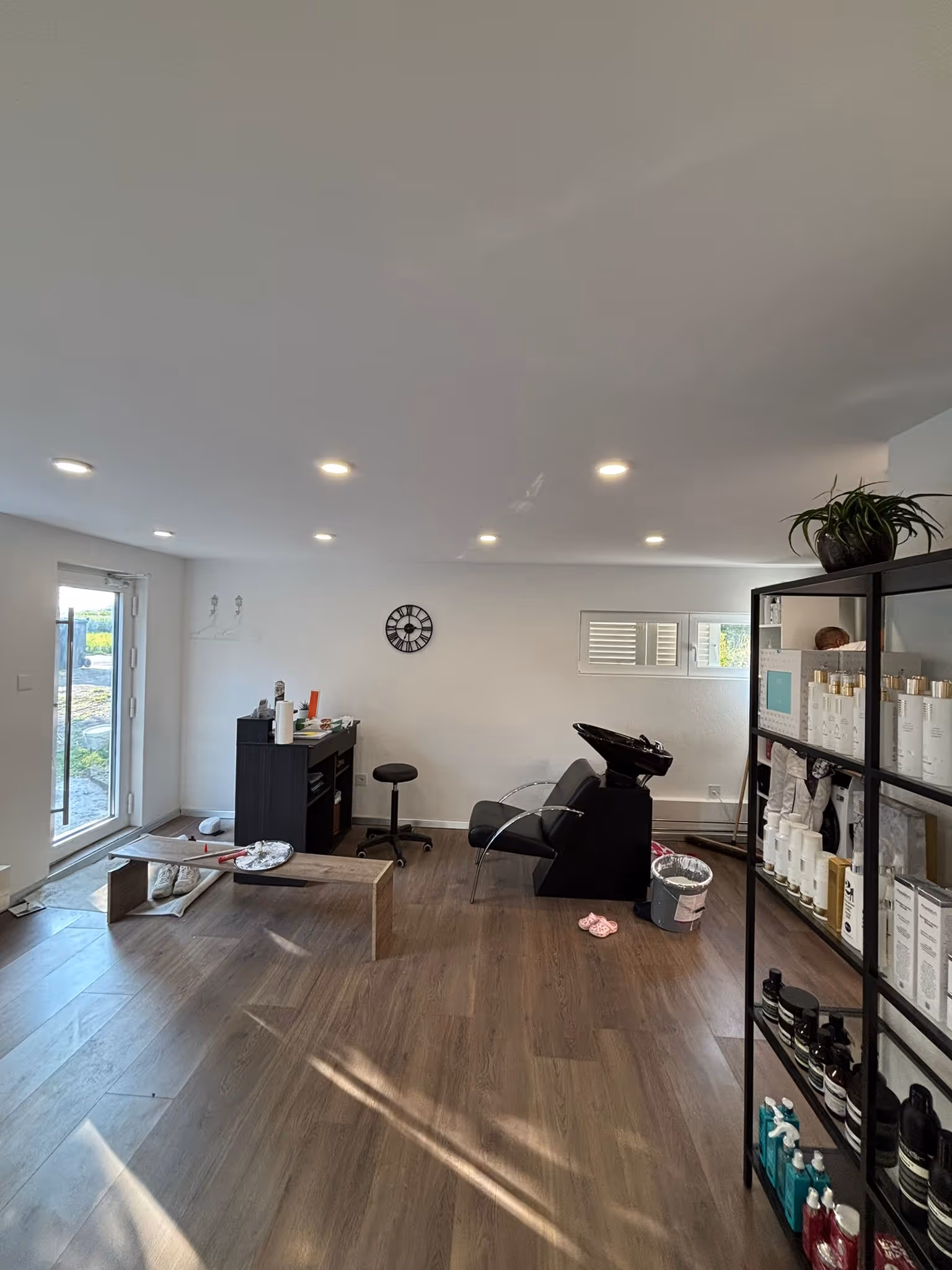 Heller Friseursalon mit Waschstation, Holzbank, schwarzem Regal mit Haarpflegeprodukten, Holzboden und grossem Fenster.