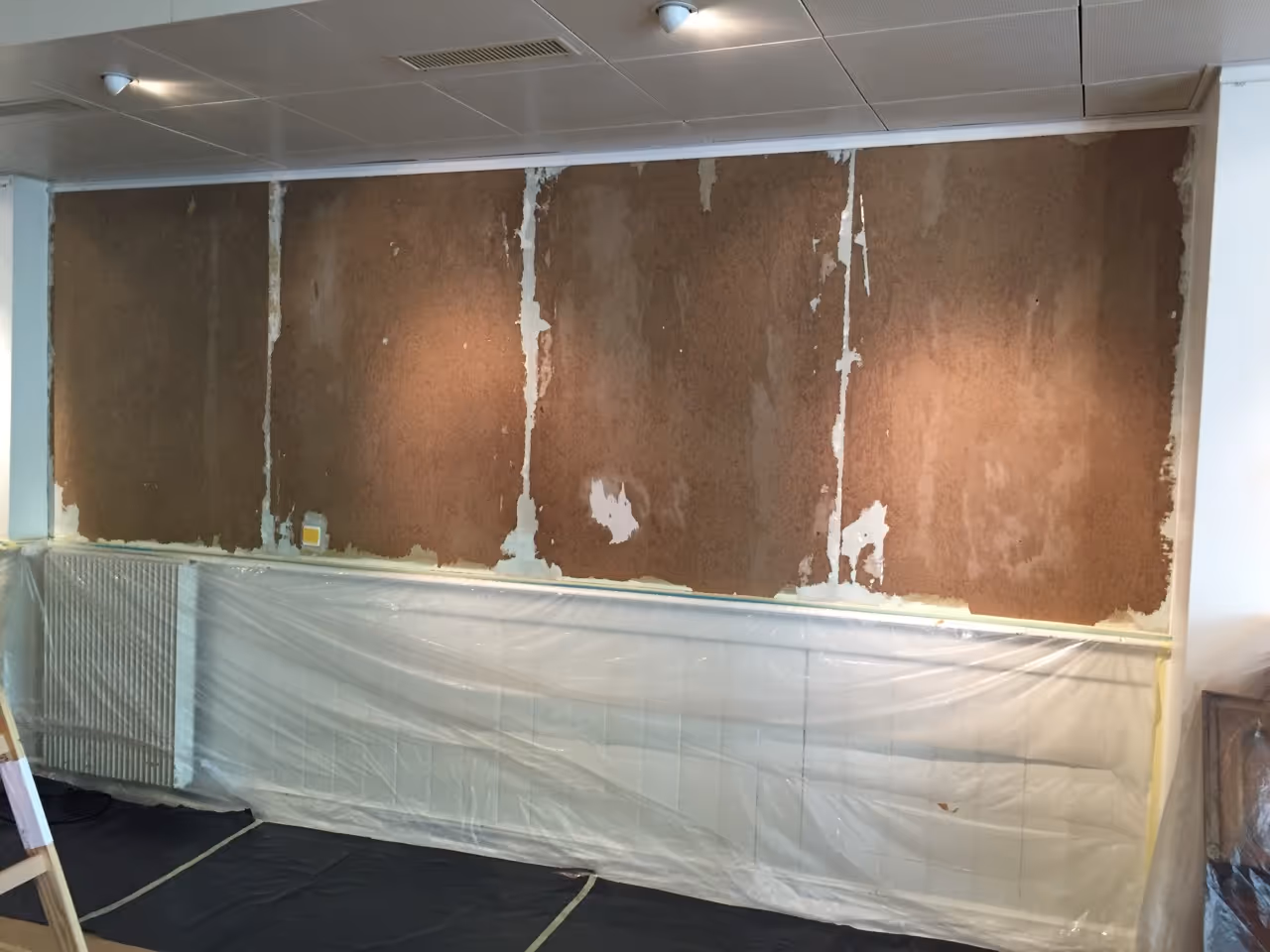 Wand mit teilweisem Putzabbruch, darunter mit Folie abgedeckte Heizkörper und Boden mit schwarzer Schutzfolie, Decke mit weißen Paneelen.