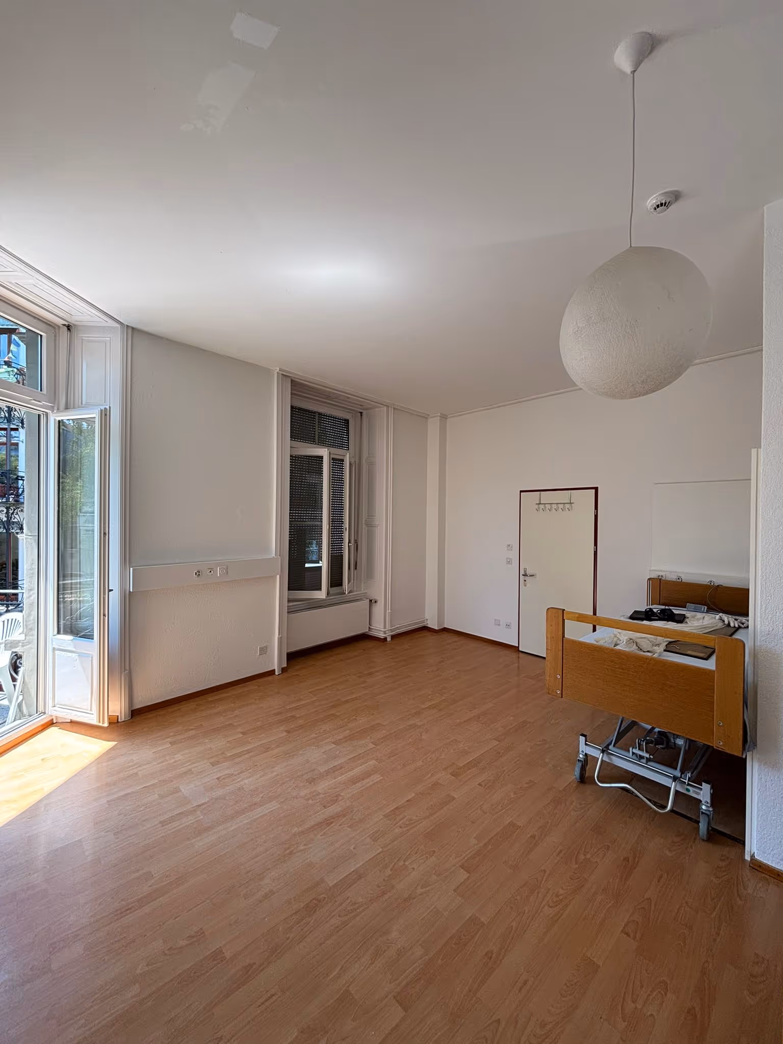 Leerer Raum mit hellen Wänden, Holzlaminatboden, offenem Fenster mit Blick auf Balkon und einem Pflegebett an der Wand.