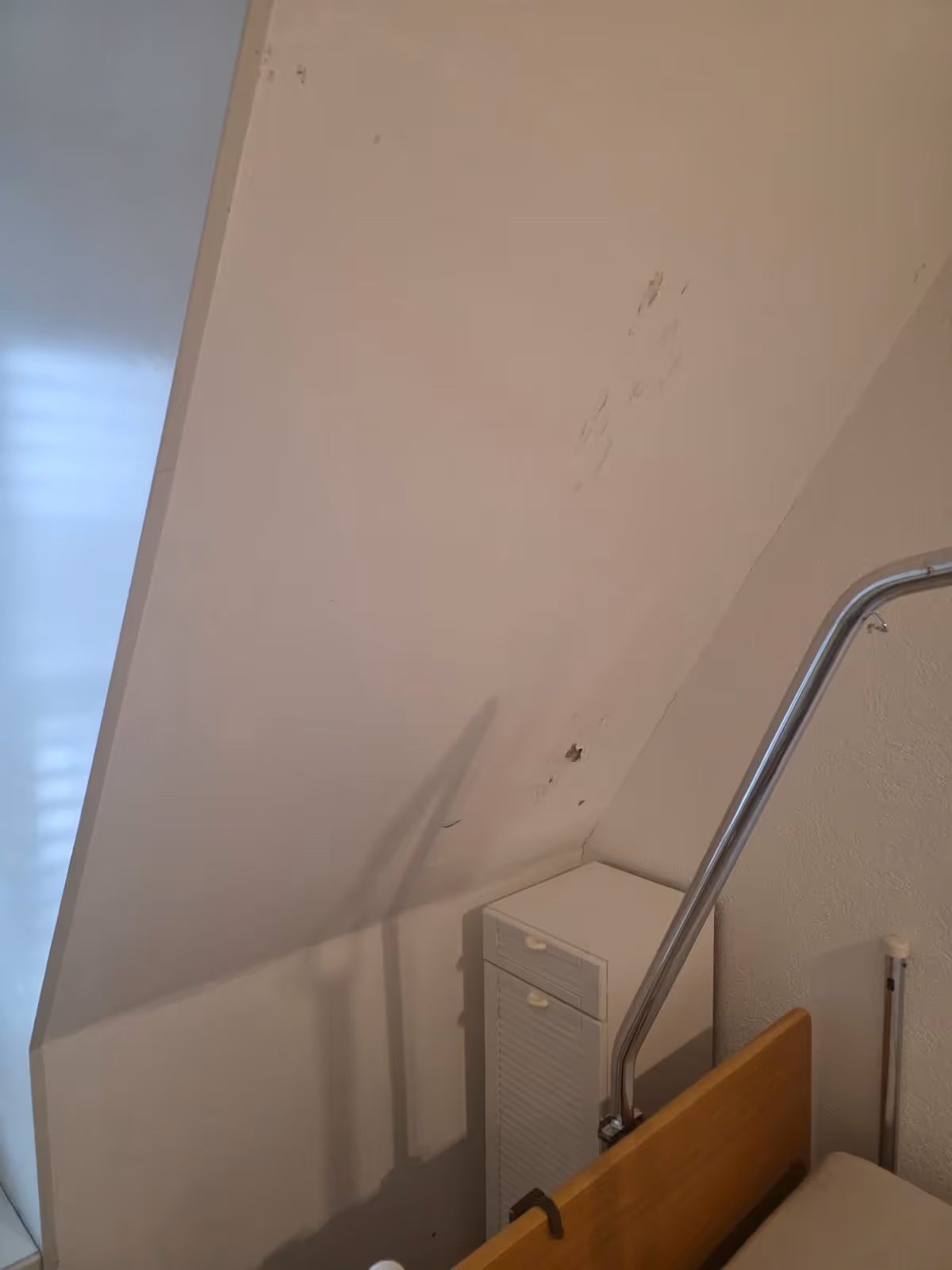 Ecke eines Zimmers mit schräger Decke, einem Bett mit Holzrahmen und Metallgeländer sowie einem kleinen weißen Nachttisch.