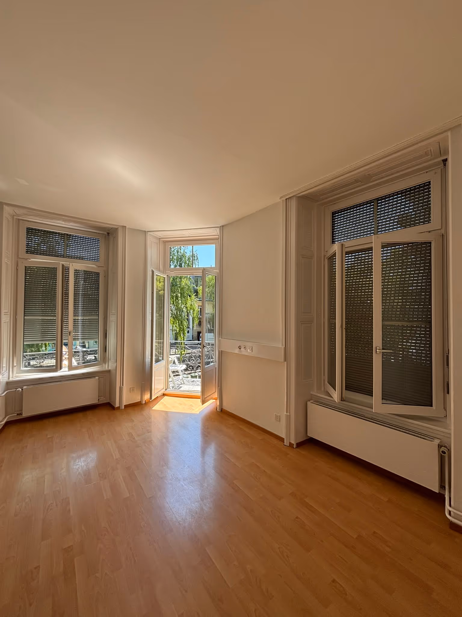 Helles, leeres Zimmer mit Holzparkett, zwei Fenstern mit Jalousien und einer offenen Glastür zu einem Balkon mit Blick auf Bäume und ein Gebäude.
