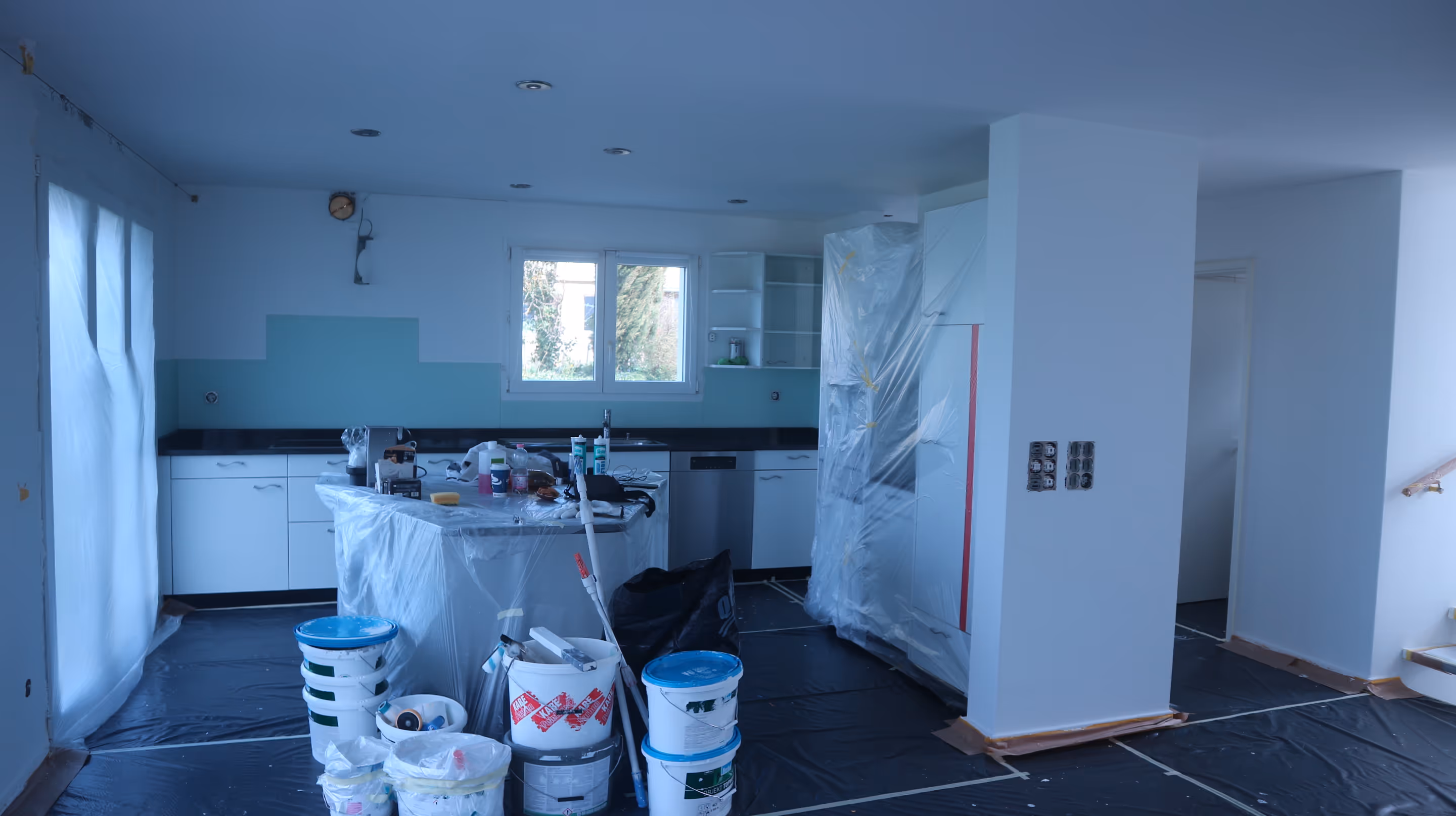 Küche in Renovierung mit abgedeckter Arbeitsfläche, Farbeimern und Schutzfolie auf Boden und Schränken.