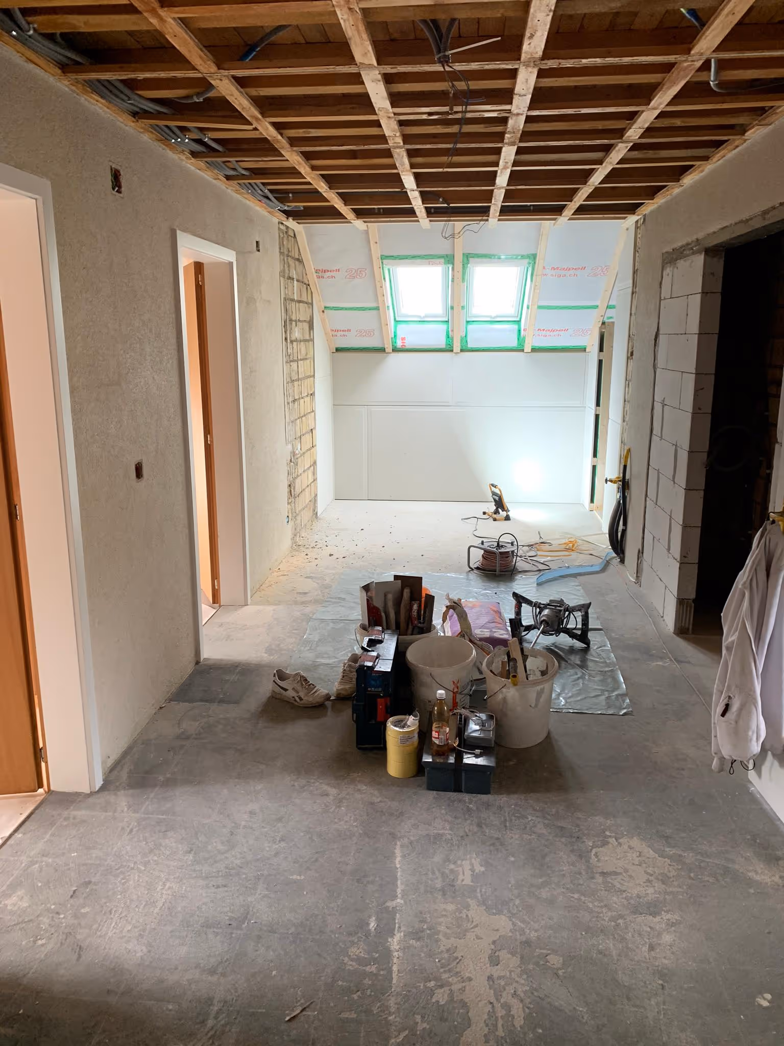 Innenraum einer Baustelle mit freigelegter Decke, unvollendeten Wänden und Baustellenwerkzeugen auf dem Boden.