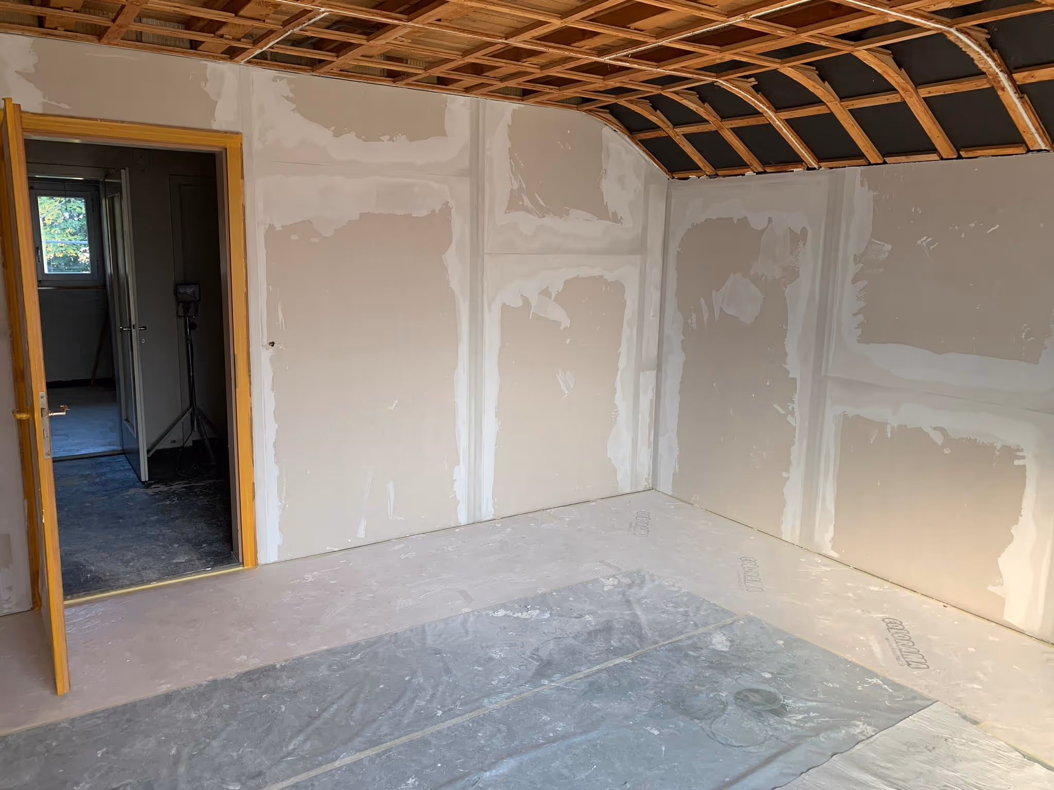 Innenraum in einem unfertigen Zustand mit verputzten Wänden, sichtbaren Holzlatten an der Decke und offenem Türrahmen in einen angrenzenden Raum.