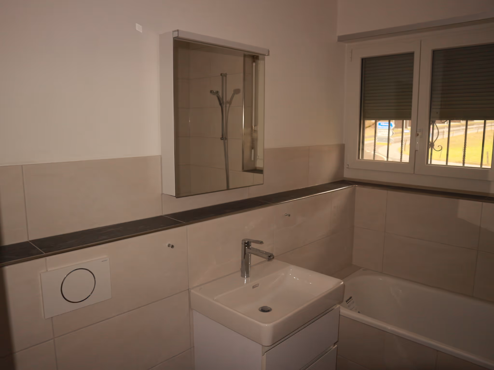 Modernes Badezimmer mit weissem Waschbecken, Unterbauschrank, eingebauter Toilette und Badewanne unter einem Fenster mit Rollläden.