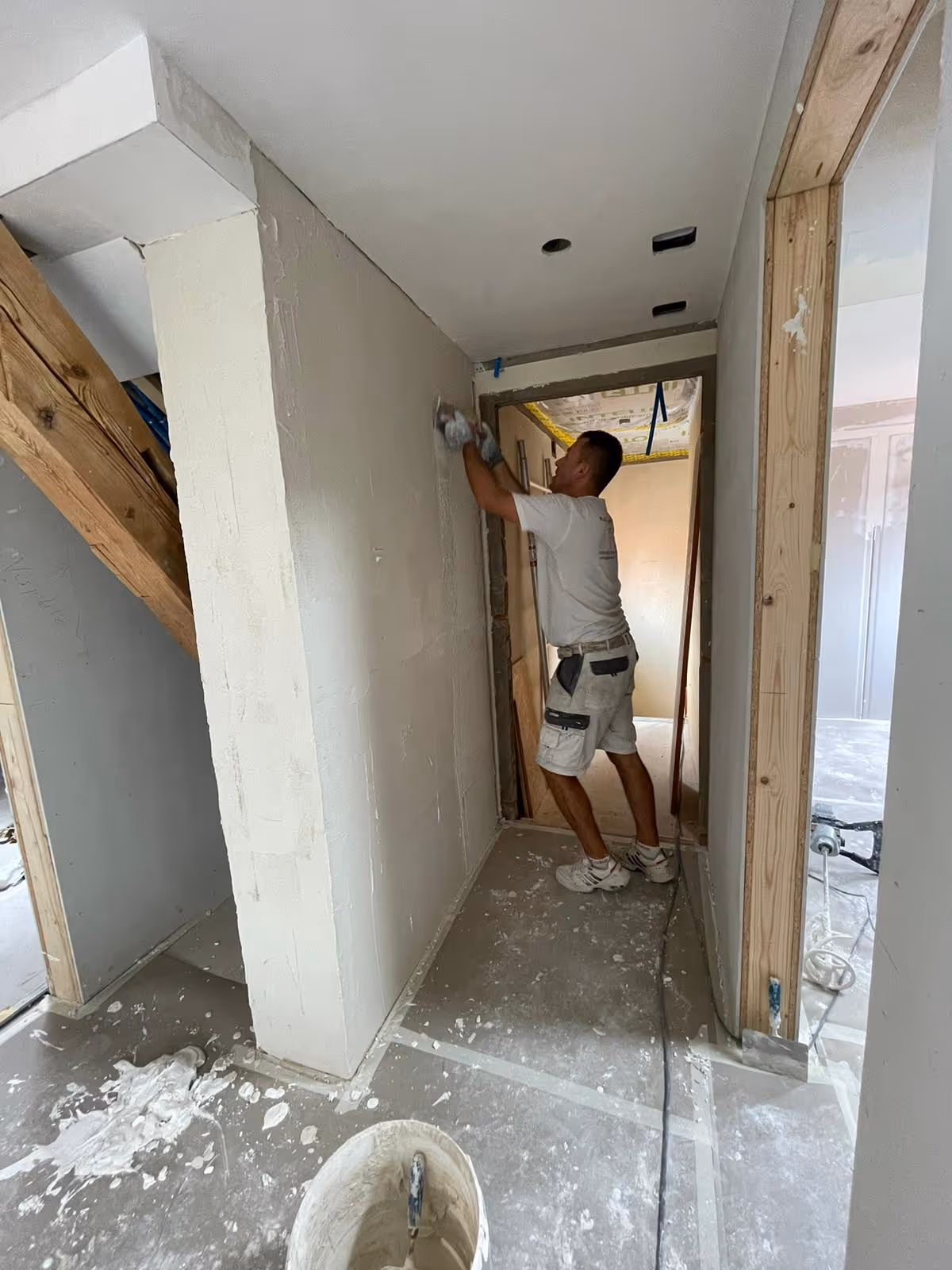Handwerker trägt weiße Kleidung und verputzt eine Wand in einem Raum unter Renovierung.