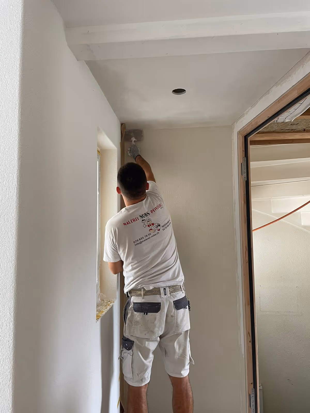 Maler trägt weisse Kleidung und streicht mit einer Kelle die obere Ecke einer Wand in einem renovierten Raum.
