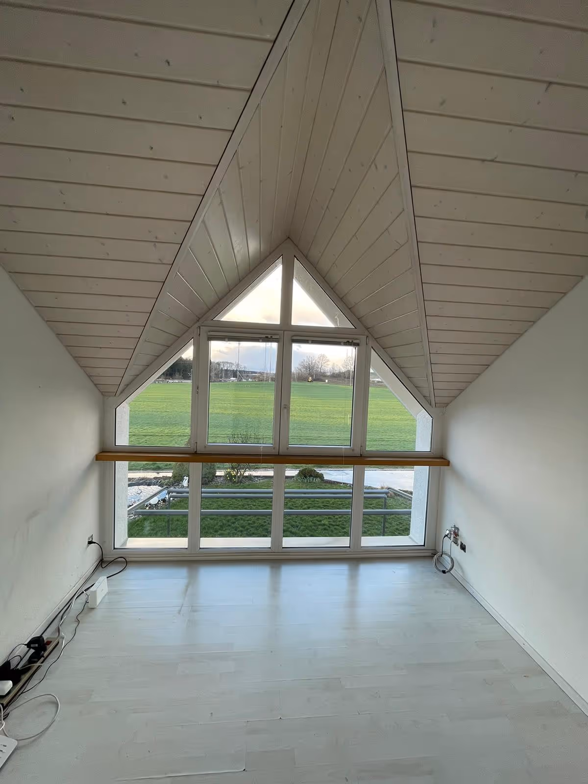 Leerer Raum mit schräger Holzdecke und großen Fensterfronten mit Blick auf eine grüne Wiese.