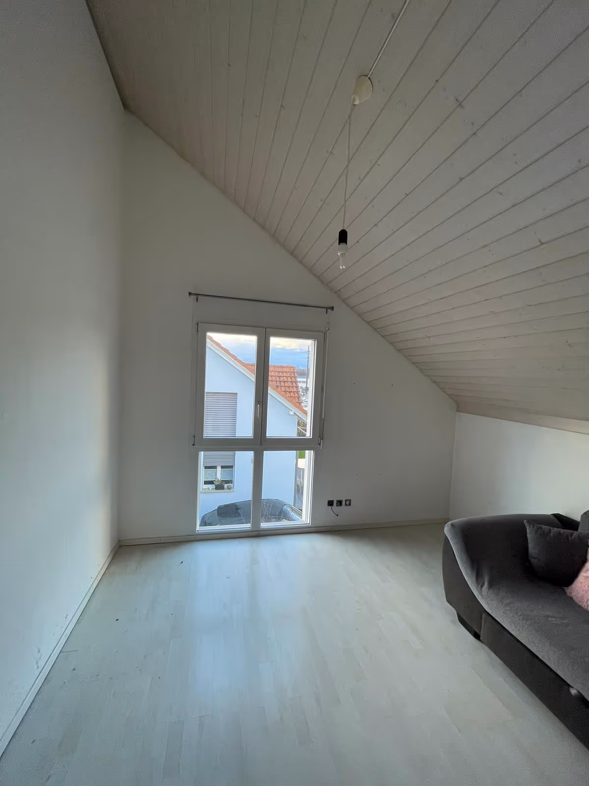 Leerer Raum mit schräger Decke, Fenster mit Blick auf ein Nachbarhaus und einem grauen Sofa mit rosa Kissen rechts.