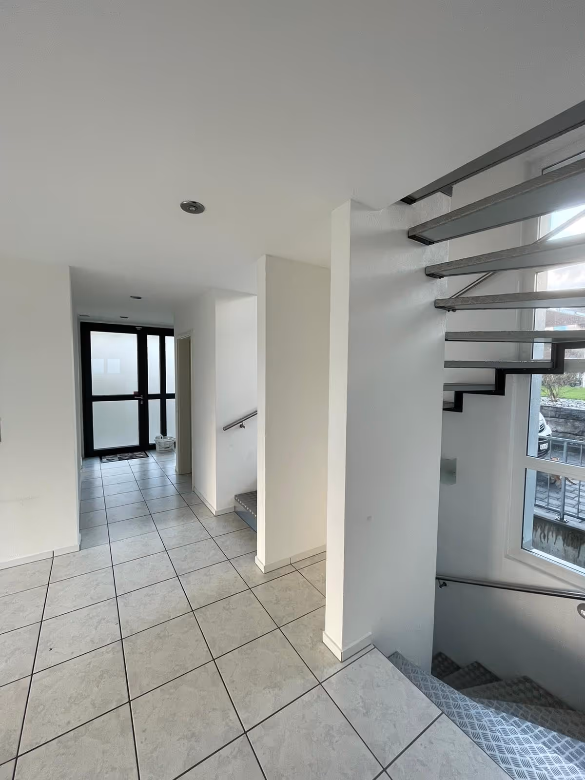 Moderner heller Eingangsbereich mit weiss gefliestem Boden, Treppenhaus und Glastür mit Milchglasscheiben.