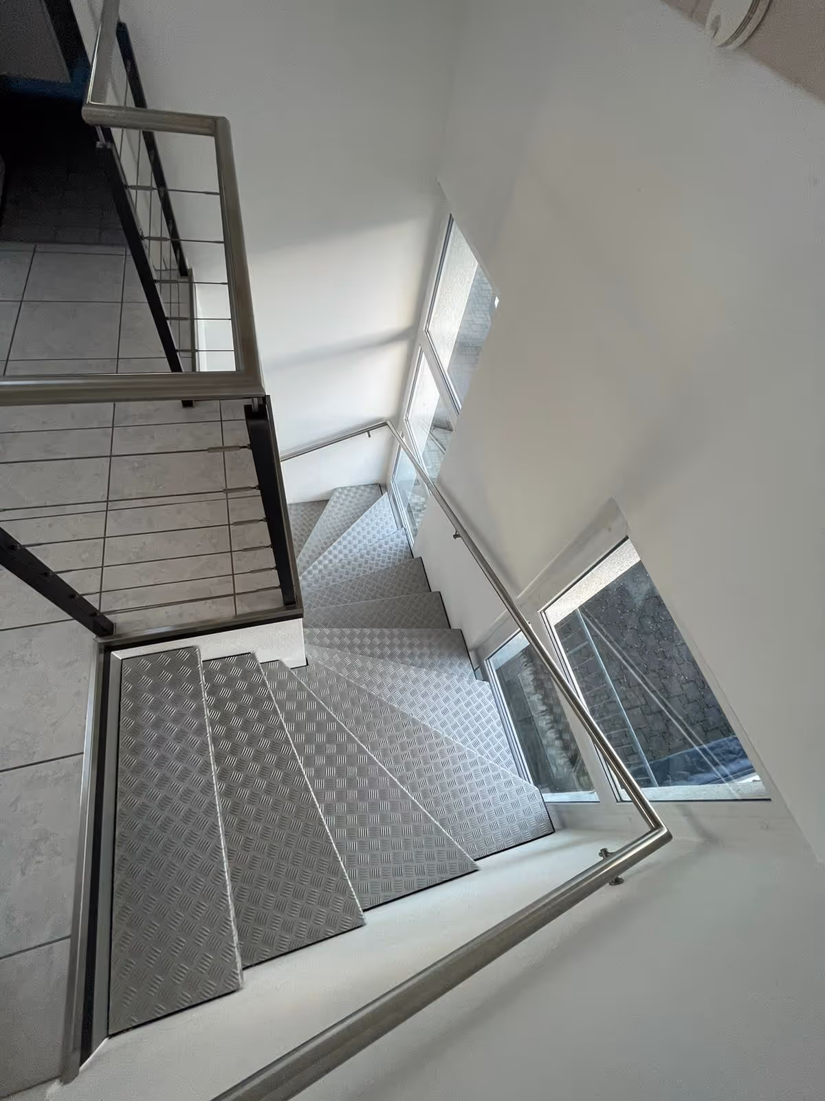 Moderner Treppenabgang mit grauen rutschfesten Metallstufen und Edelstahlgeländer neben grossen Fenstern.