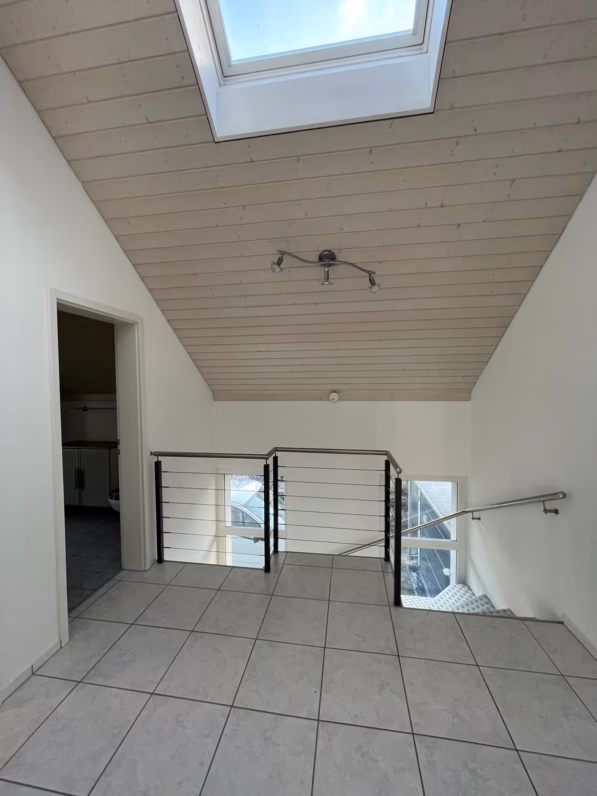 Helle, moderne Galerie mit grauen Bodenfliesen, Holzdeckenbalken und Treppe mit Edelstahlgeländer unter einem Dachfenster.