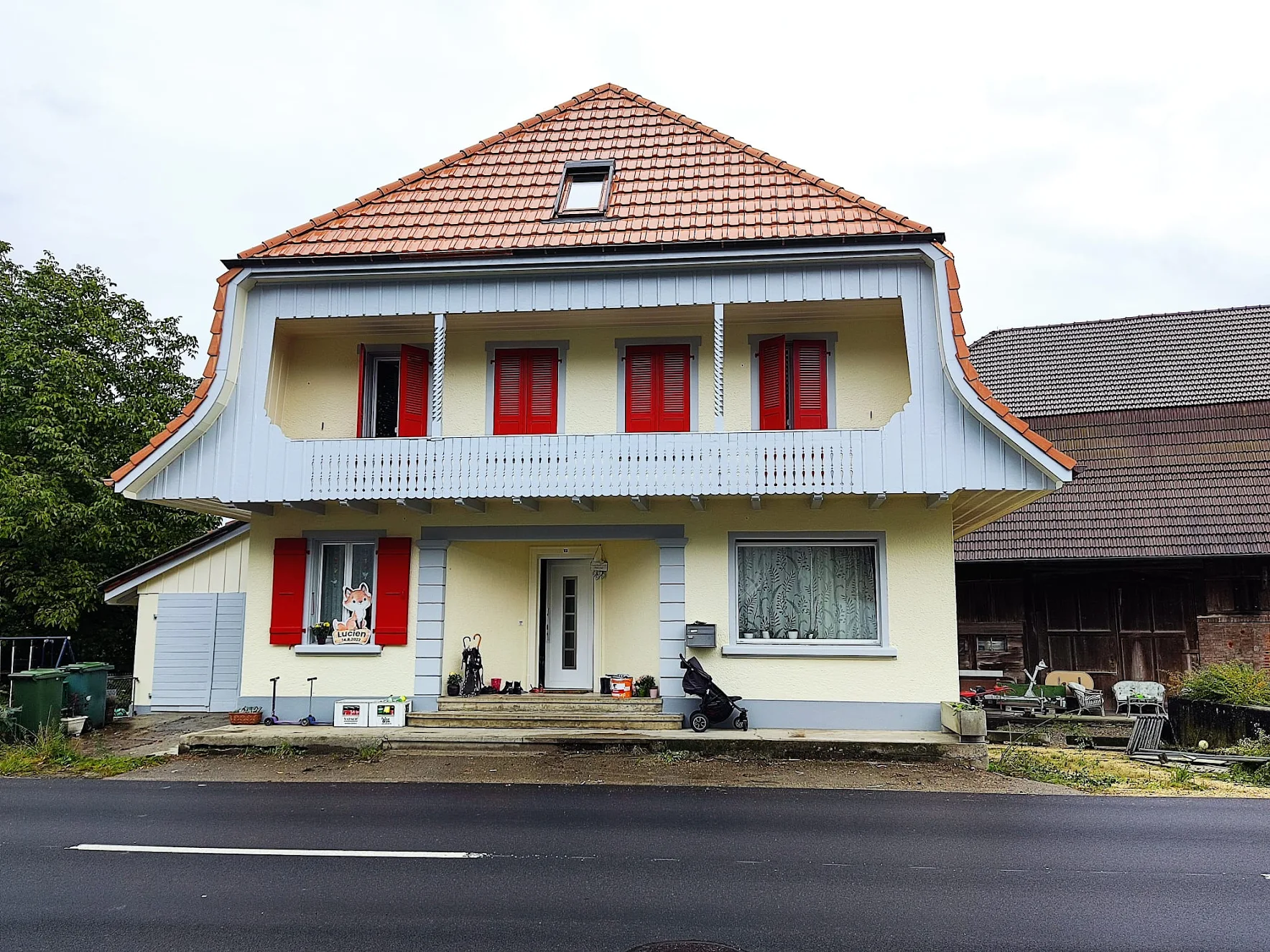 Zweistöckiges Haus mit gelber Fassade, rotem Schindeldach und roten Fensterläden, davor Straße und Gartenutensilien.