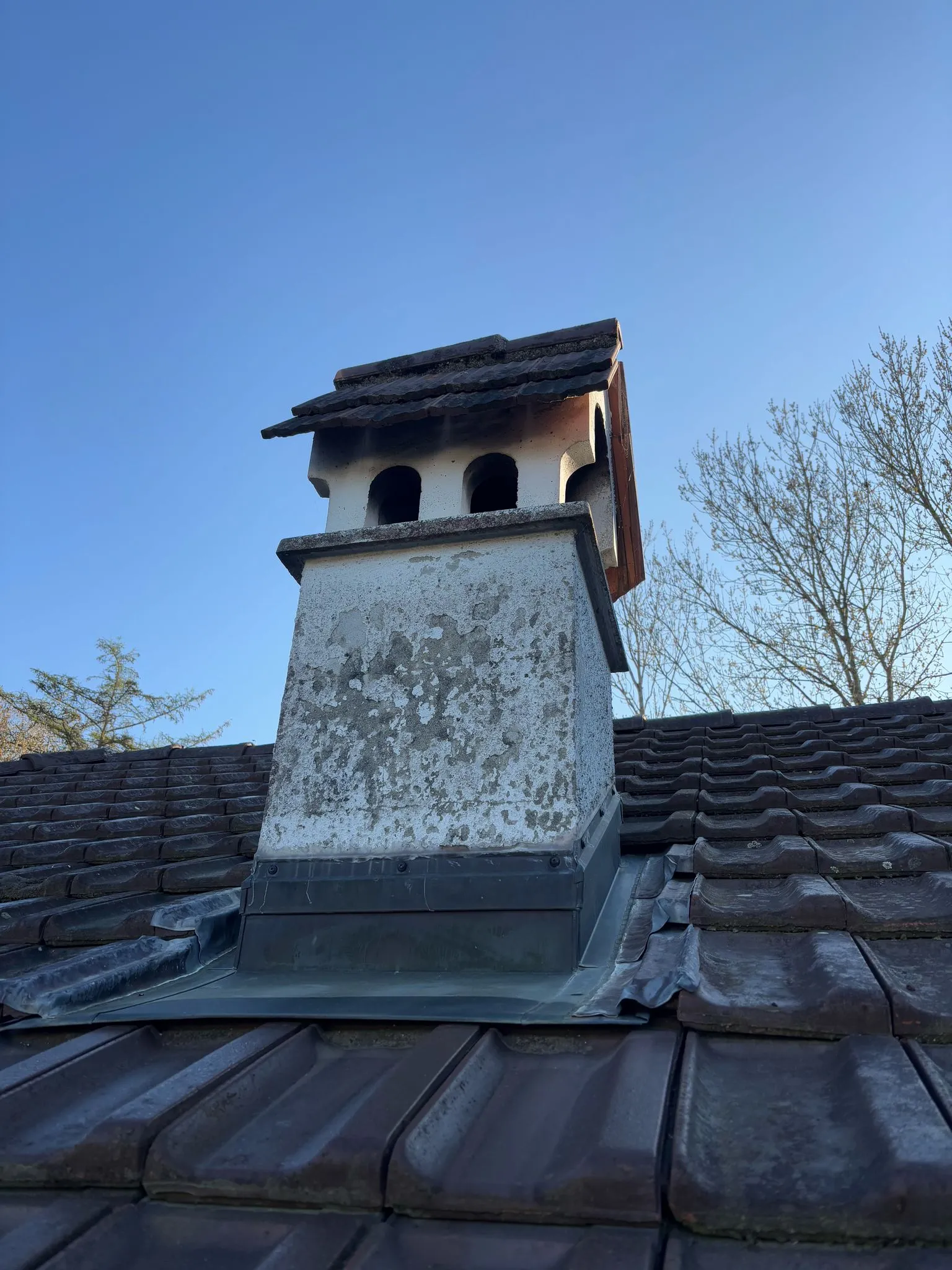 Schornstein mit dunklen Rußspuren auf einem Ziegeldach vor klarem blauem Himmel und kahlen Bäumen.
