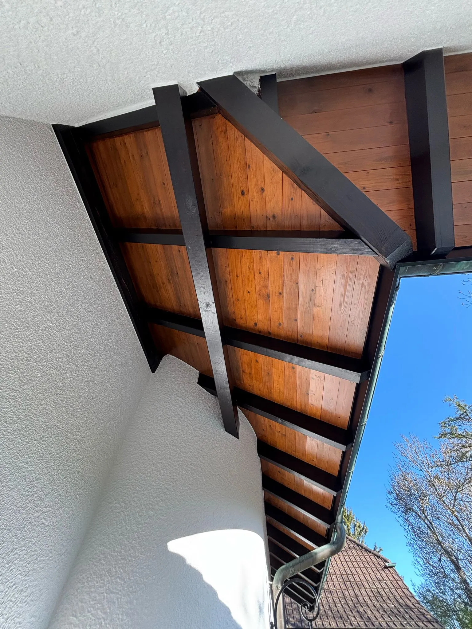 Blick auf ein Dach mit dunklen Holzbalken und Holzverkleidung unter dem Dachüberstand eines weissen Hauses.