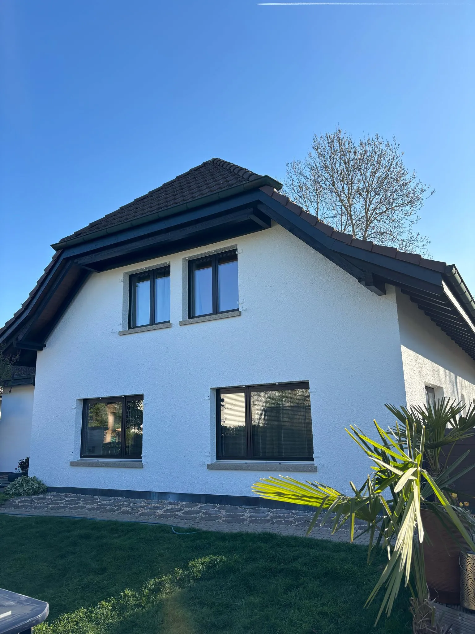 Ein weisses Haus mit dunklem Ziegeldach, vier Fenstern und einem Baum im Hintergrund unter klarem blauem Himmel.
