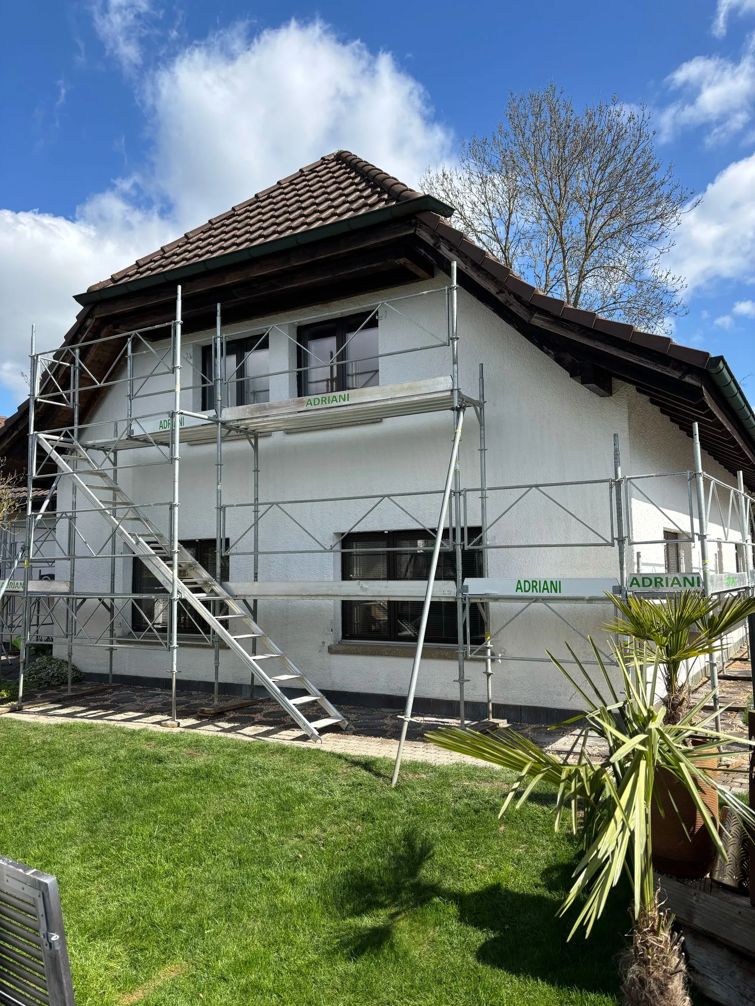 Haus mit braunem Ziegeldach und weißer Fassade, eingerüstet mit Baugerüst bei sonnigem Himmel.