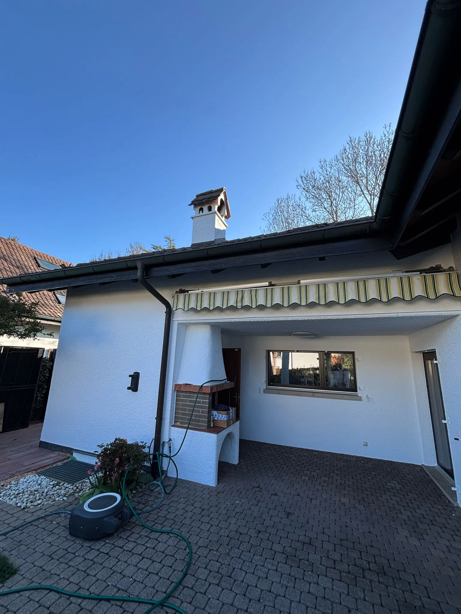 Weisses Haus mit offener Terrasse, Markise und einer Aussenküche mit Schornstein unter blauem Himmel.