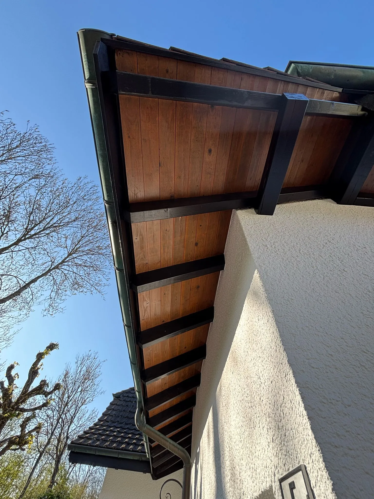 Untersicht des Holzdachs mit dunklen Balken und heller Putzfassade eines Hauses bei blauem Himmel.