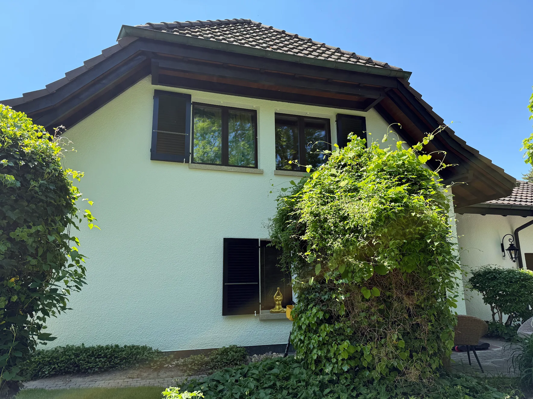 Haus mit weisser Fassade, braunem Ziegeldach und schwarzen Fensterläden, umgeben von grünem Buschwerk und Pflanzen.
