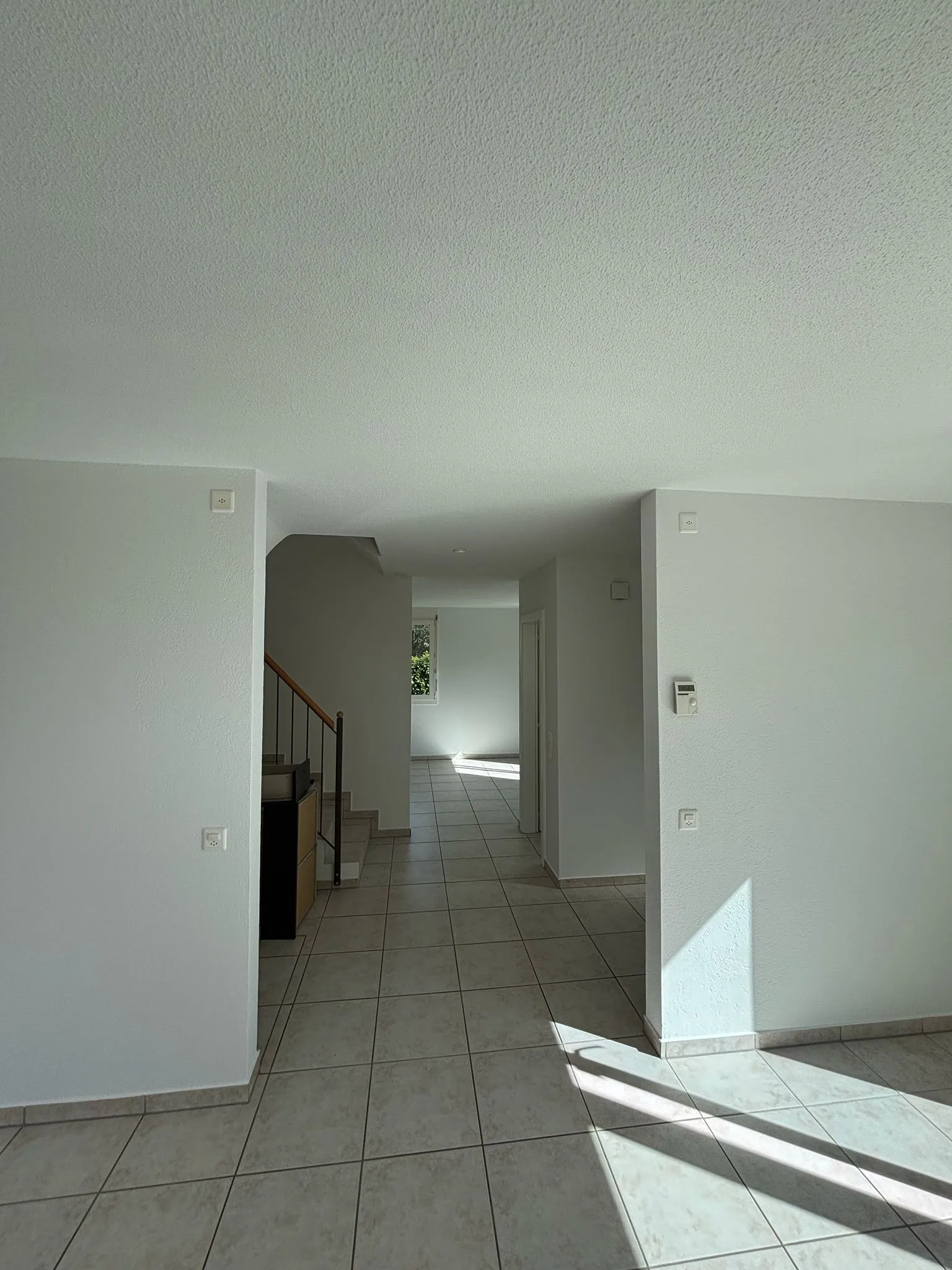 Leerer heller Raum mit weissen Wänden, hellen Fliesen und Treppe mit Holz- und Metallgeländer.