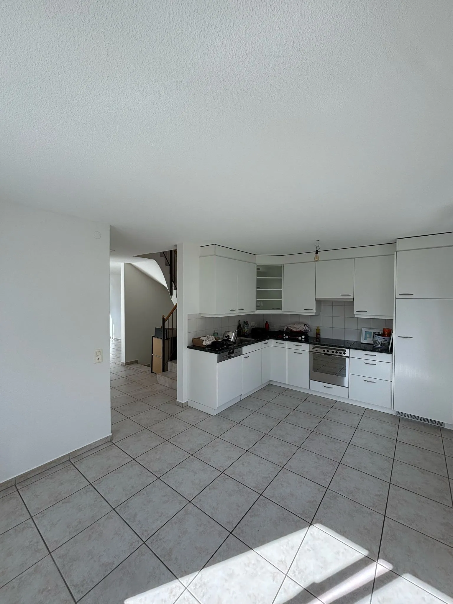 Moderne Küche mit weissen Schränken, grauen Fliesenboden und Treppe im Hintergrund.