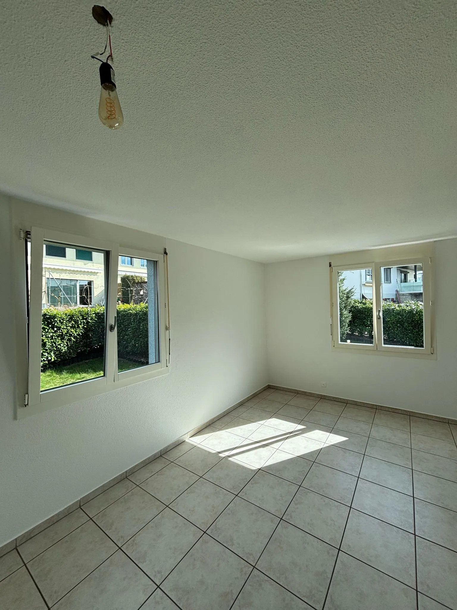 Leerer Raum mit zwei geöffneten Fenstern, weiß gestrichenen Wänden, hellgrauen Bodenfliesen und einer einzelnen hängenden Glühbirne an der Decke.