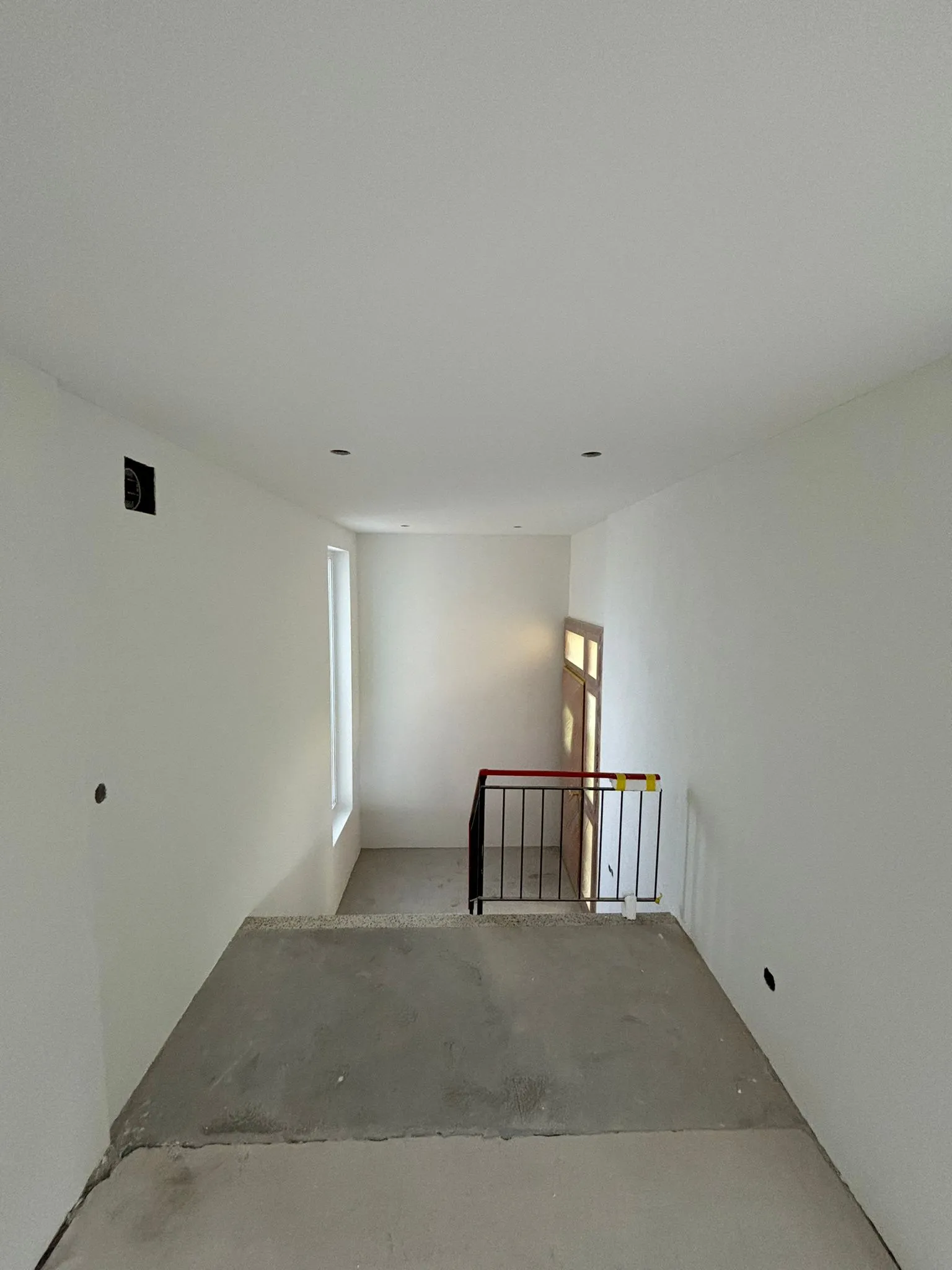 Innenraum eines unfertigen Treppenhauses mit weißen Wänden, rotem Geländer und einem Fenster.