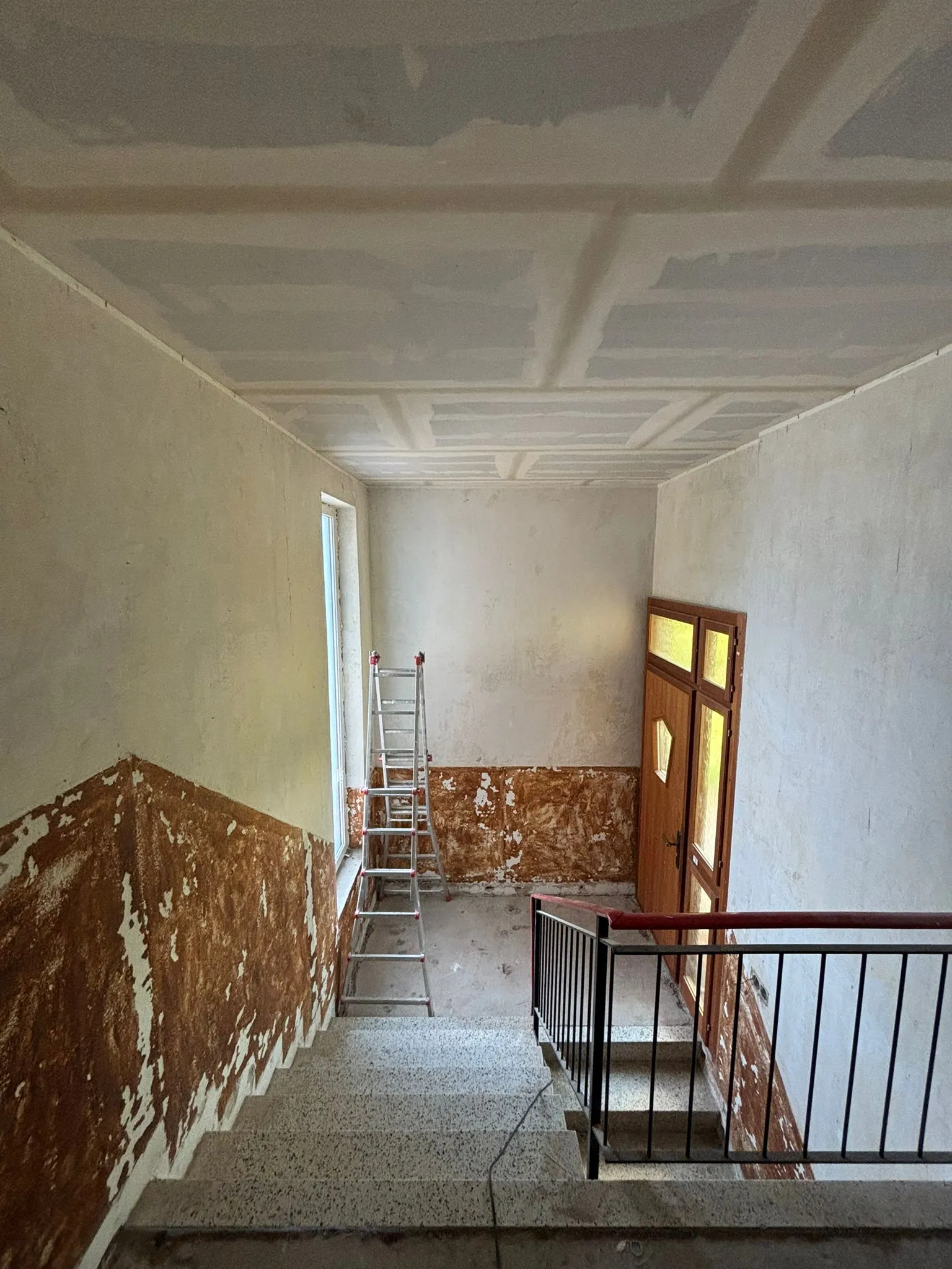 Treppenhaus in einem Gebäude unter Renovierung mit teilweise entfernten Wandputzen, einer Leiter auf der Fensterbank und einer Holztür mit gelben Fenstern.