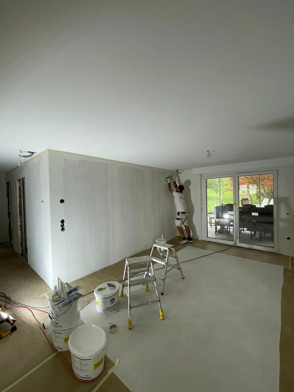 Mann bearbeitet Wand im Innenraum eines Hauses mit Rollenfarbe, umgeben von Farbeimern und Leitern, neben grossen Glasschiebetüren.