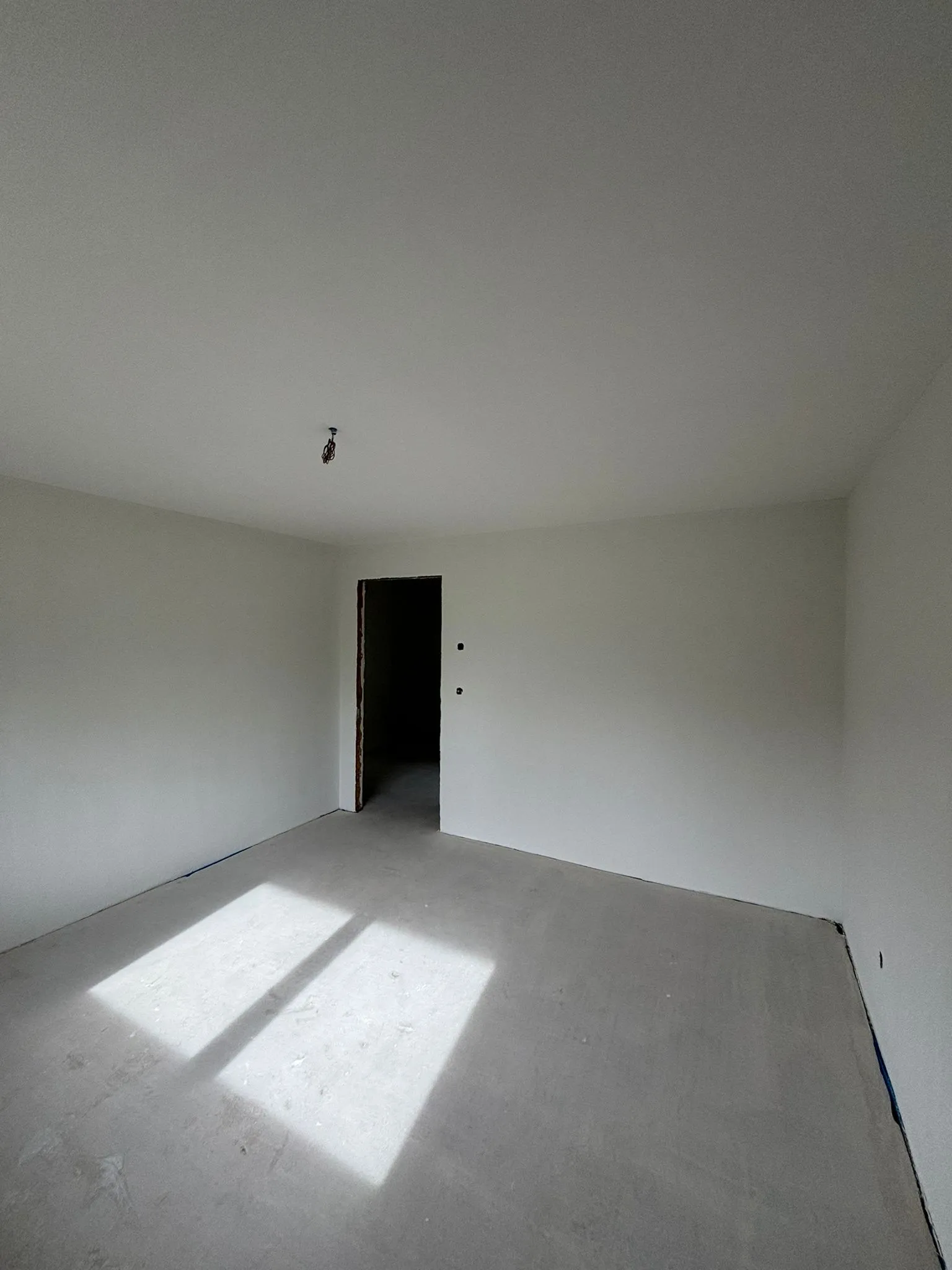 Unmöblierter, leerer Raum mit weißen Wänden und Boden, Sonnenlicht fällt durch ein nicht sichtbares Fenster auf den Boden, offener Durchgang im Hintergrund.