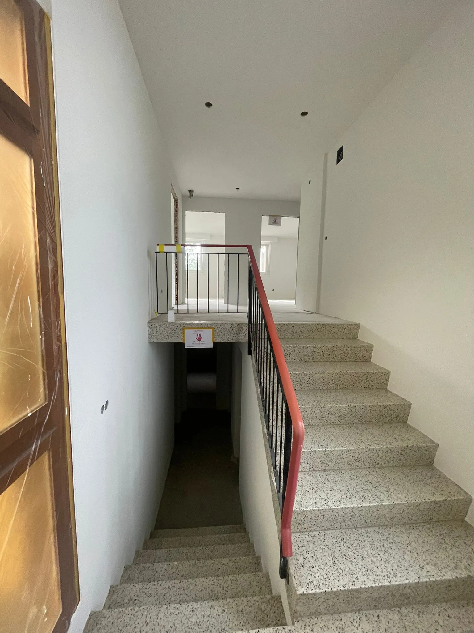 Innere Treppe in einem Gebäude mit hellen Wänden, roten Handläufen und einer Absperrung am oberen Treppenabsatz.