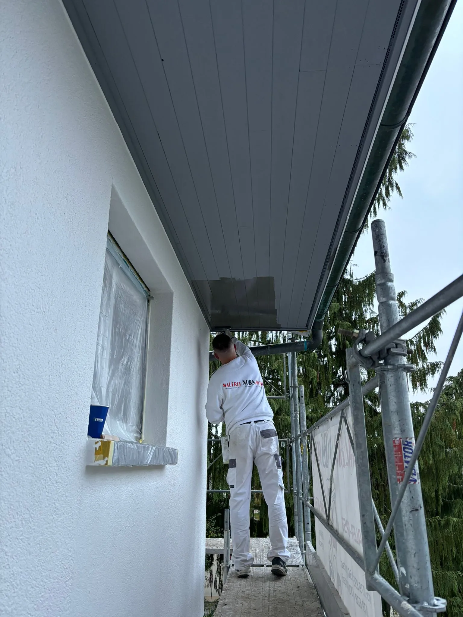 Maler steht auf einem Gerüst und streicht die Decke eines überdachten Bereichs an einem weißen Gebäude.