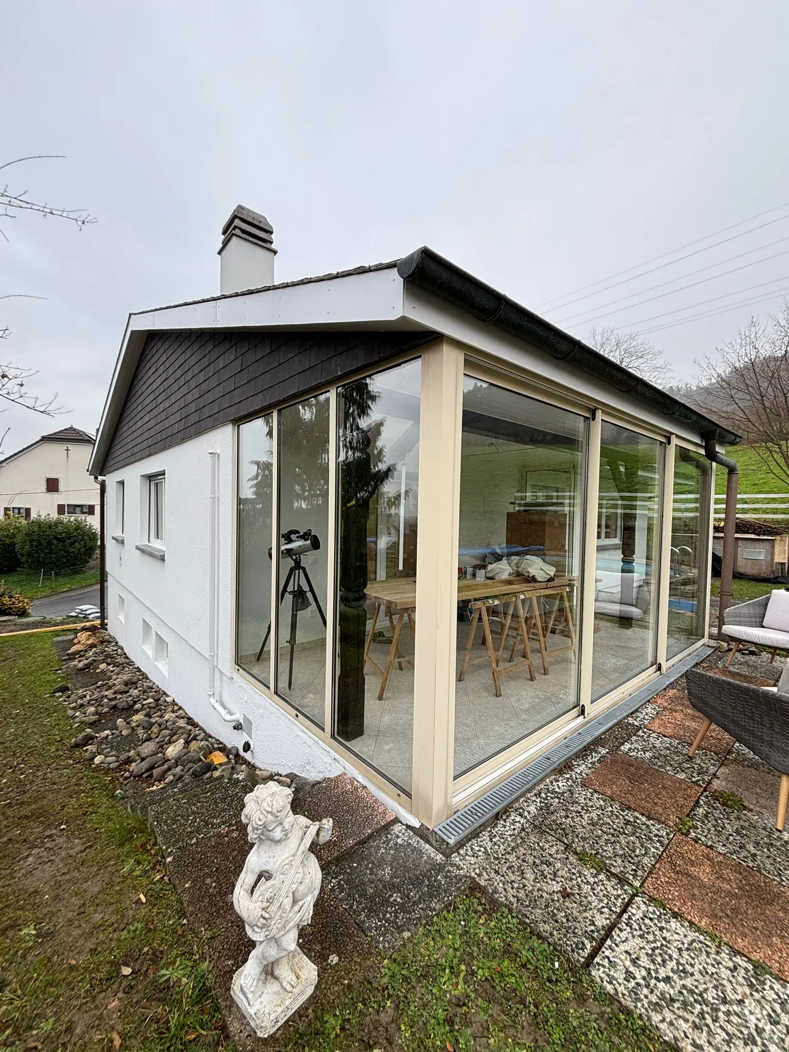 Kleines Haus mit verglaster Terrasse, Garten mit einer Engelfigur aus Stein und Außenmöbeln auf einer gepflasterten Fläche.