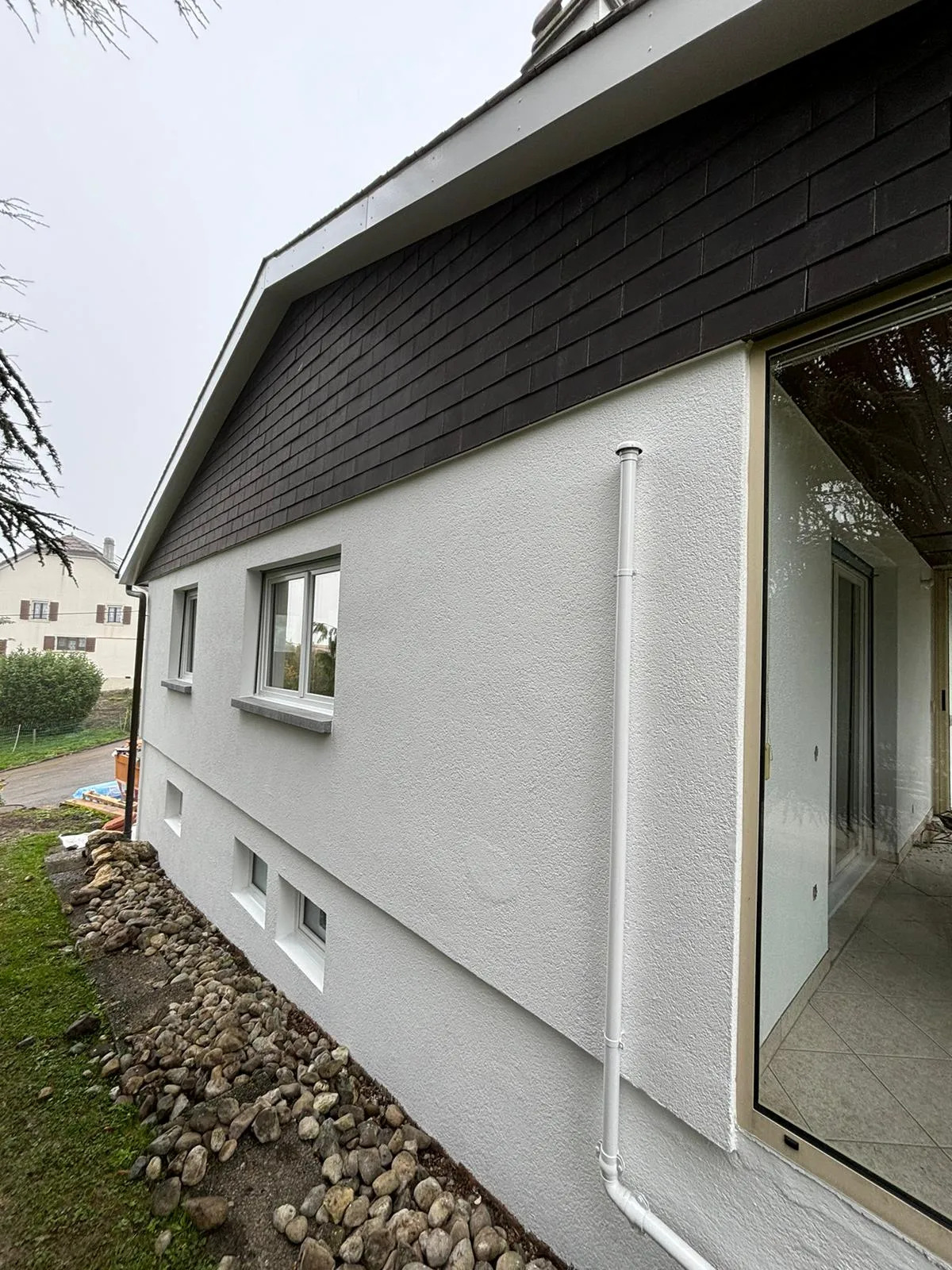 Weiße Fassade eines Einfamilienhauses mit kleinen Fenstern und schwarzen Dachziegeln, davor ein Beet mit Steinen.