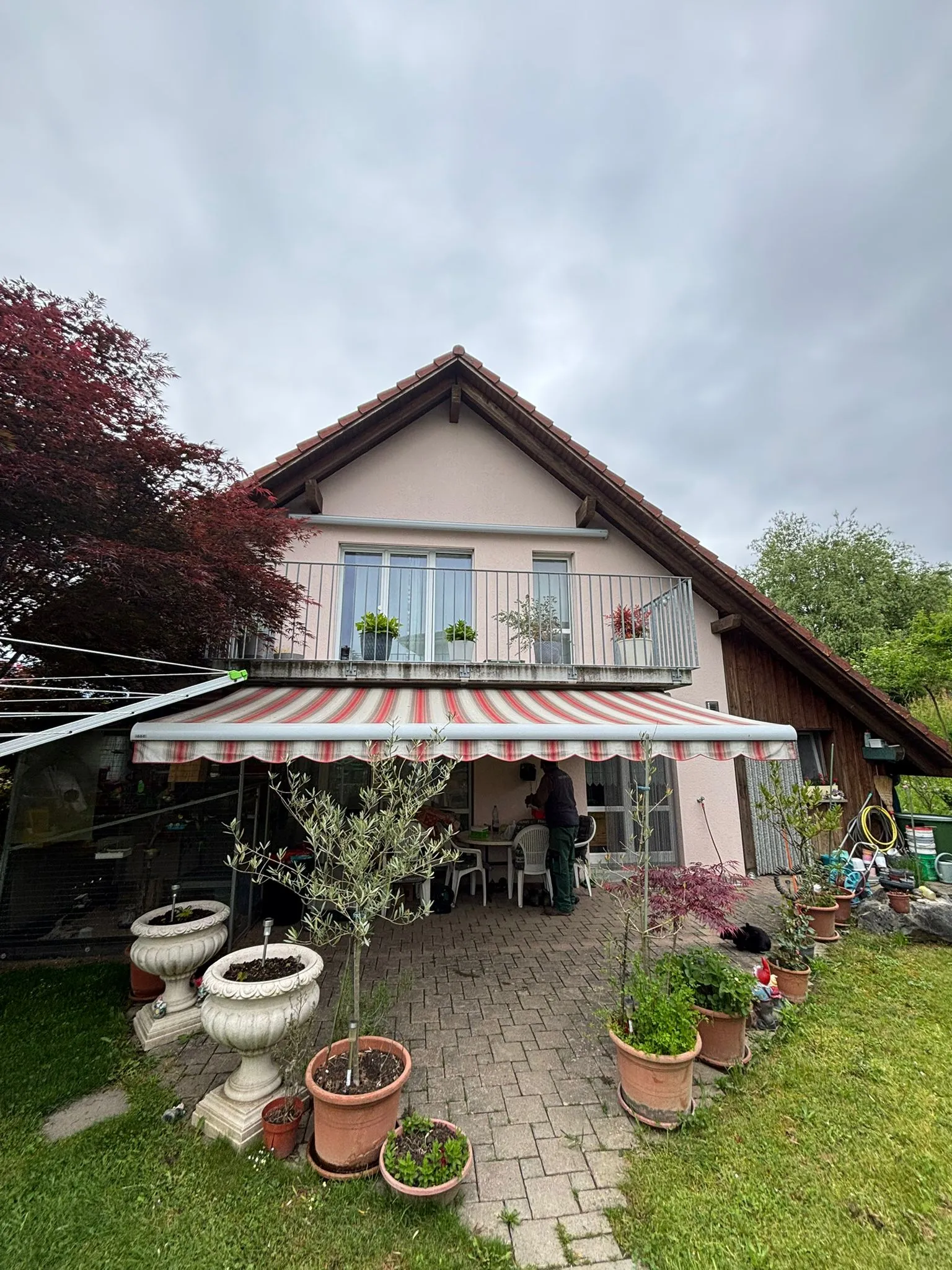 Haus mit Balkon und rot-weiß gestreiftem Markisentuch, Terrasse mit Topfpflanzen und Person, die an einem Tisch steht.