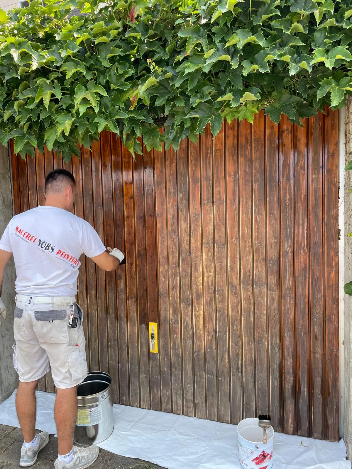 Mann streicht eine Holztür mit Pinsel und dunkler Holzfarbe unter einem Blätterdach.