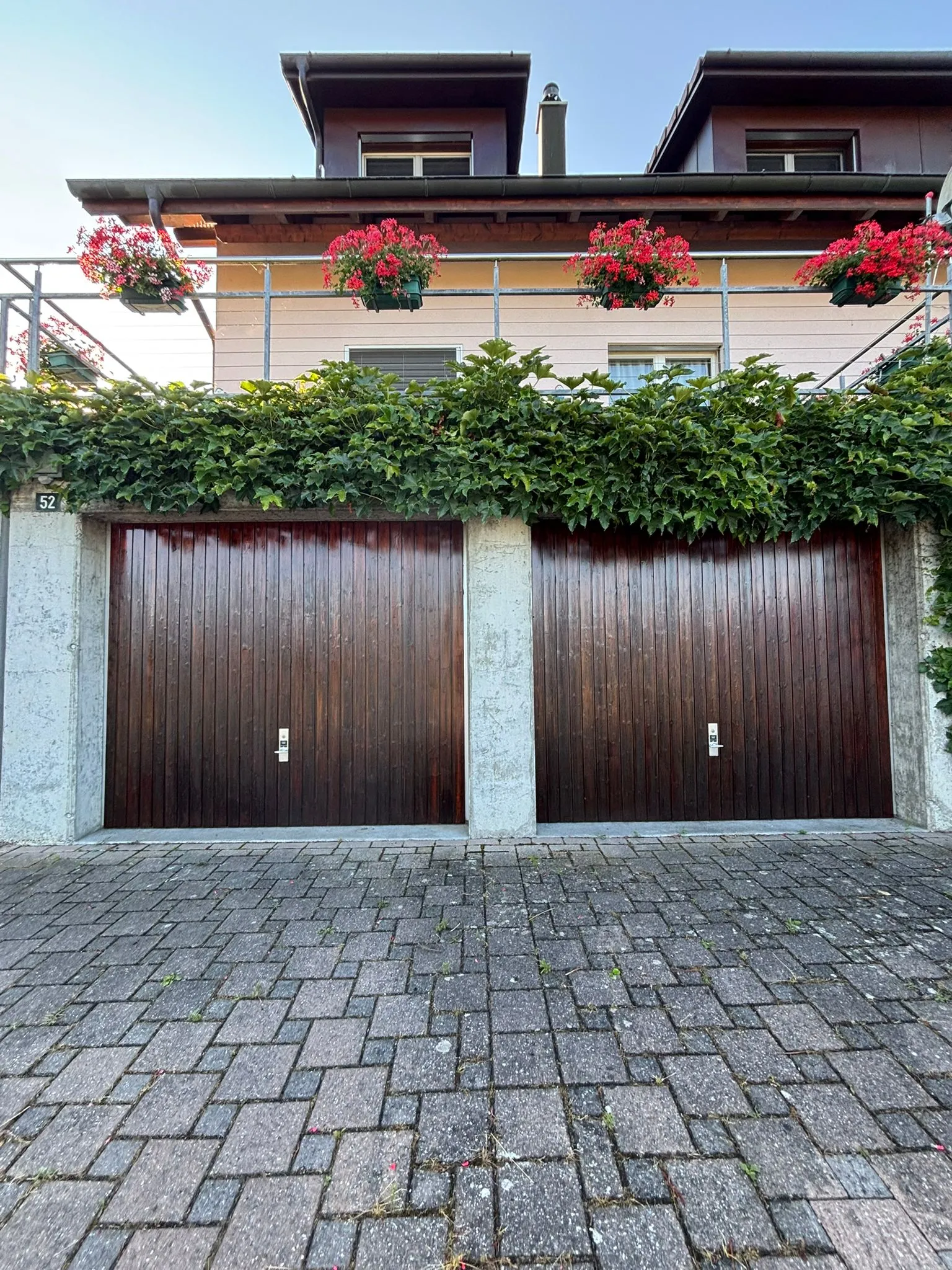 Zwei dunkle Holztore, umrahmt von Beton, mit grünen Kletterpflanzen darüber und roten Blumen in Blumenkästen auf dem Balkon eines Wohnhauses.