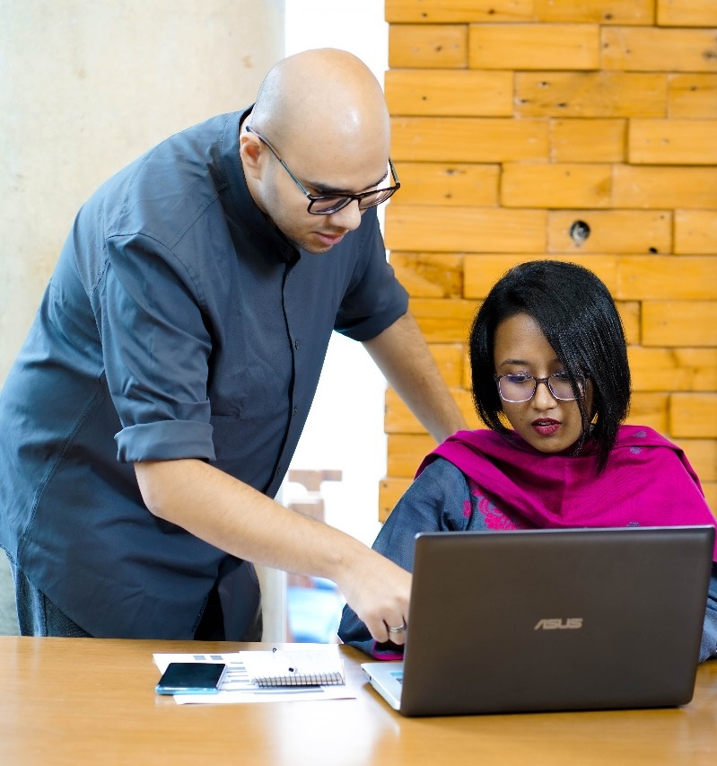 Man pointing at laptop screen helping woman in pink shawl seated at wooden desk with coffee mug.