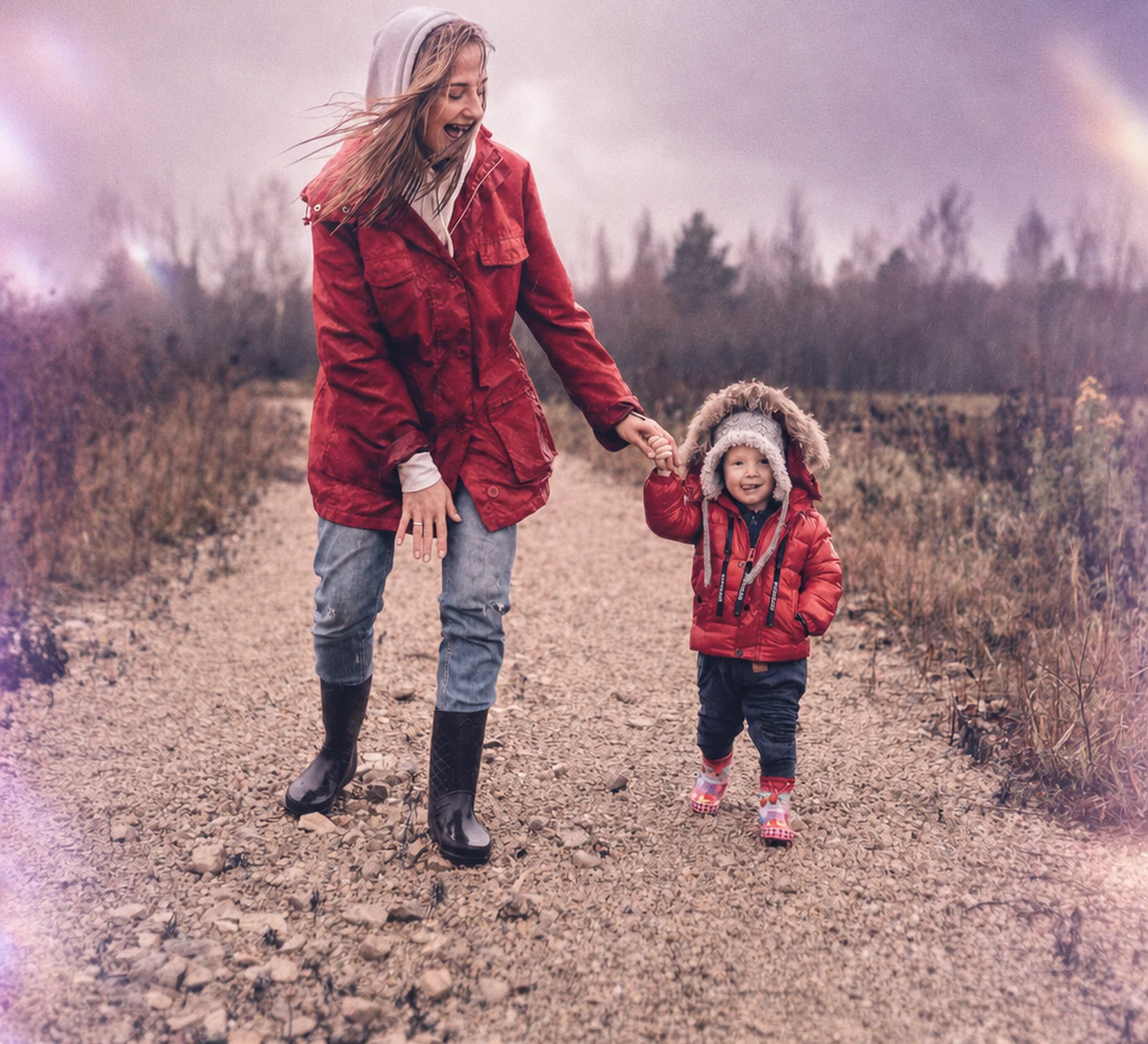 A mother and her toddler in matching red coats walk hand in hand along a gravel country path
