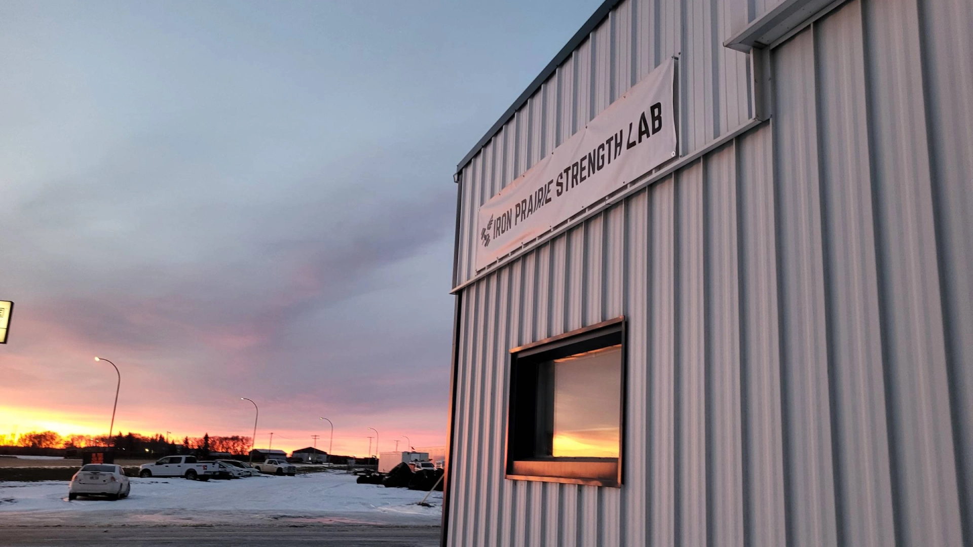 The sun sets behind Iron Prairie Strength Lab in Moosomin, SK, with a sign visible in the foreground.